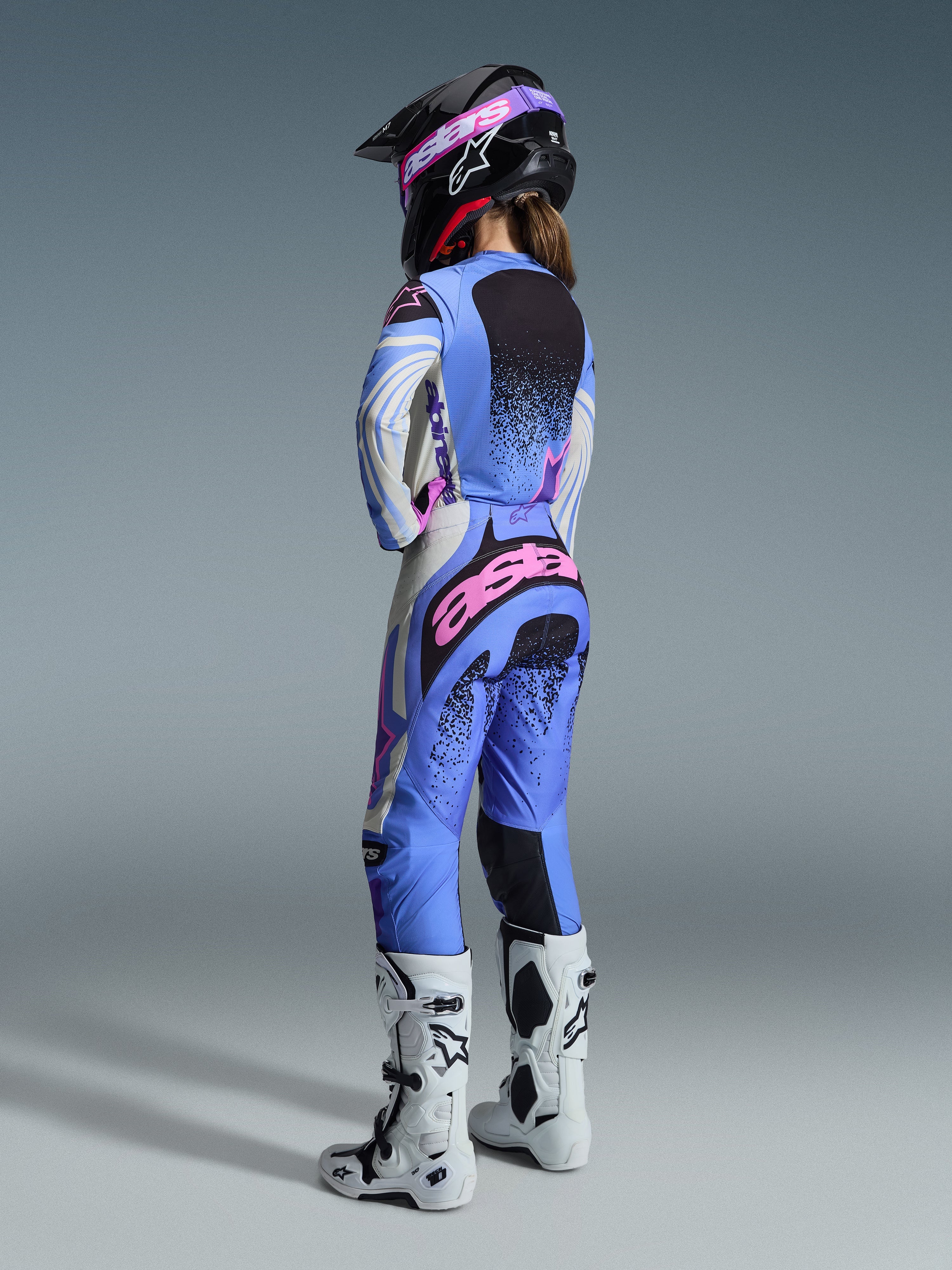 Alpinestars 2026 Stella Techstar Nomur Gear Set, vue arrière d'une pilote de motocross portant un ensemble maillot et pantalon bleu clair, blanc et rose avec des détails mouchetés noirs, assorti à un casque noir et des bottes blanches, équipement de course professionnel pour femmes.