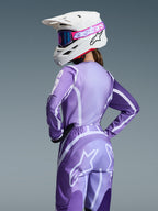 Motorcrossrijder in Alpinestars 2026 Stella Fluid Apex Gear Set, paarse en lavendelkleurige jersey en broek met witte graphics, witte helm met roze en paarse crossbril, specifieke damespasvorm, gecoördineerde uitrusting voor off-road racen