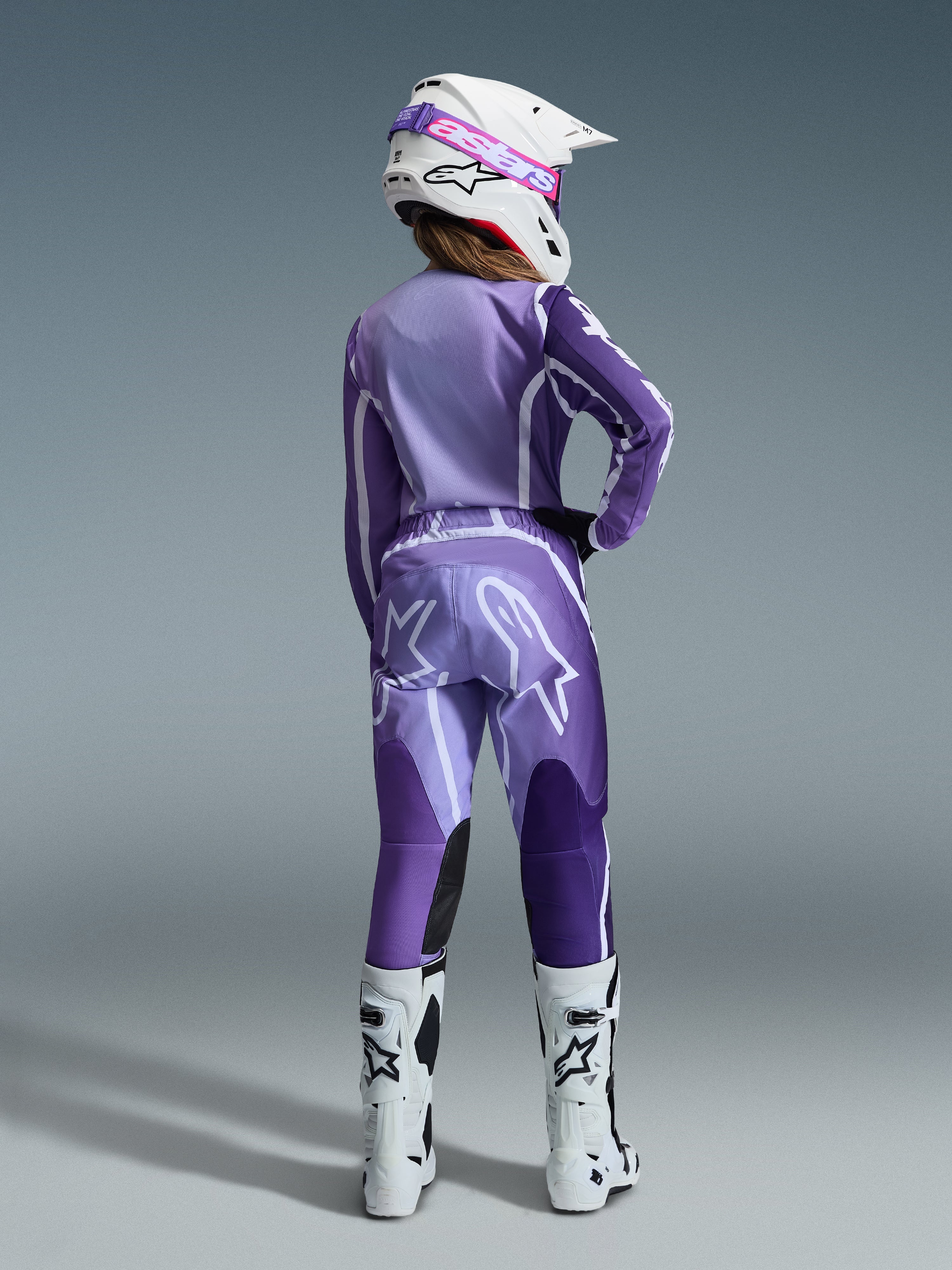 Alpinestars 2026 Stella Fluid Apex Gear Set, motocrosskleding voor dames, paars en lavendel met witte accenten, vrouwelijke rijder van achteren gezien in bijpassende jersey en broek, witte helm met roze crossbril en witte laarzen, gecoördineerde sportieve race-outfit