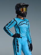 Ensemble Stella Fluid Apex Femme 2026 Alpinestars, bleu vif et noir, maillot et pantalon de motocross avec logos noirs, porté avec un casque noir, des lunettes à effet miroir doré et des gants noirs, coupe spécifique femme pour la course tout-terrain