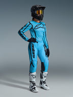 Ensemble Alpinestars Stella Fluid Apex Femme 2026 en Tropical Black, comprenant un maillot bleu clair et un pantalon assorti avec des détails noirs, porté par une pilote avec un casque noir, des lunettes irisées et des bottes blanches, équipement professionnel de motocross.