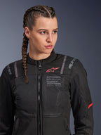 Veste Alpinestars Stella ST-2 Air, veste de moto pour femme en noir, avec panneaux en mesh pour la circulation de l'air, marquage « Alpinestars Exploration Equipment » avec coordonnées, logo rouge sur la poitrine, portée par un mannequin aux cheveux tressés