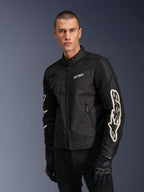 Veste Alpinestars T-Dyno Air en noir, mannequin masculin portant un blouson de moto en textile d'inspiration vintage avec panneaux en maille pour la ventilation, grands logos Alpinestars blancs sur les manches, associé à des gants de moto noirs, design d'équipement de moto urbain