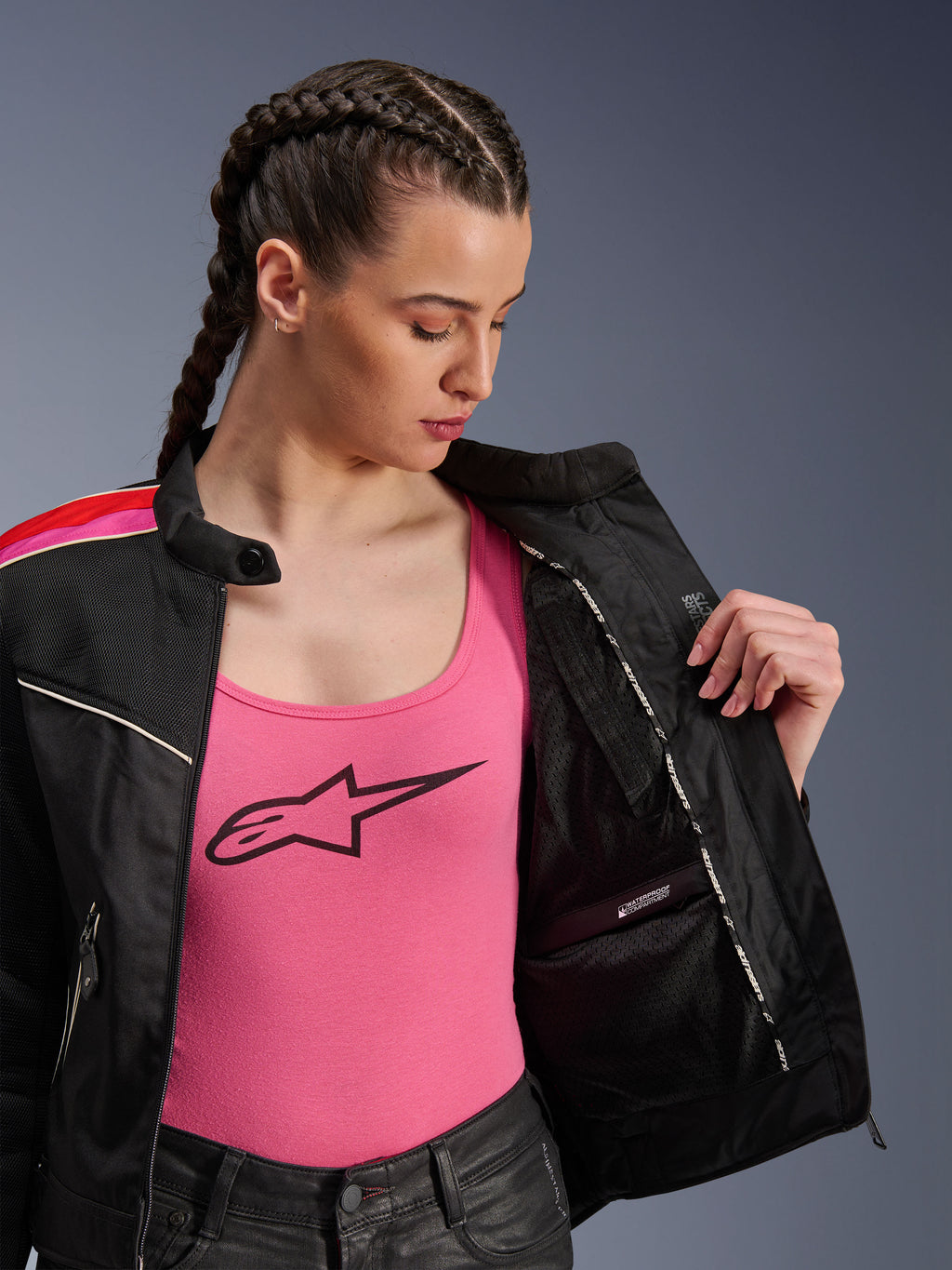 Motorradfahrerin in der Alpinestars Stella T-Dyno Air Jacke in Schwarz, Hellrot und Fuchsia, teilweise geöffnet, um das interne wasserdichte Fach zu zeigen, getragen über einem pinkfarbenen Logo-Tanktop, betont die atmungsaktiven Mesh-Einsätze und die taillierte Biker-Passform.