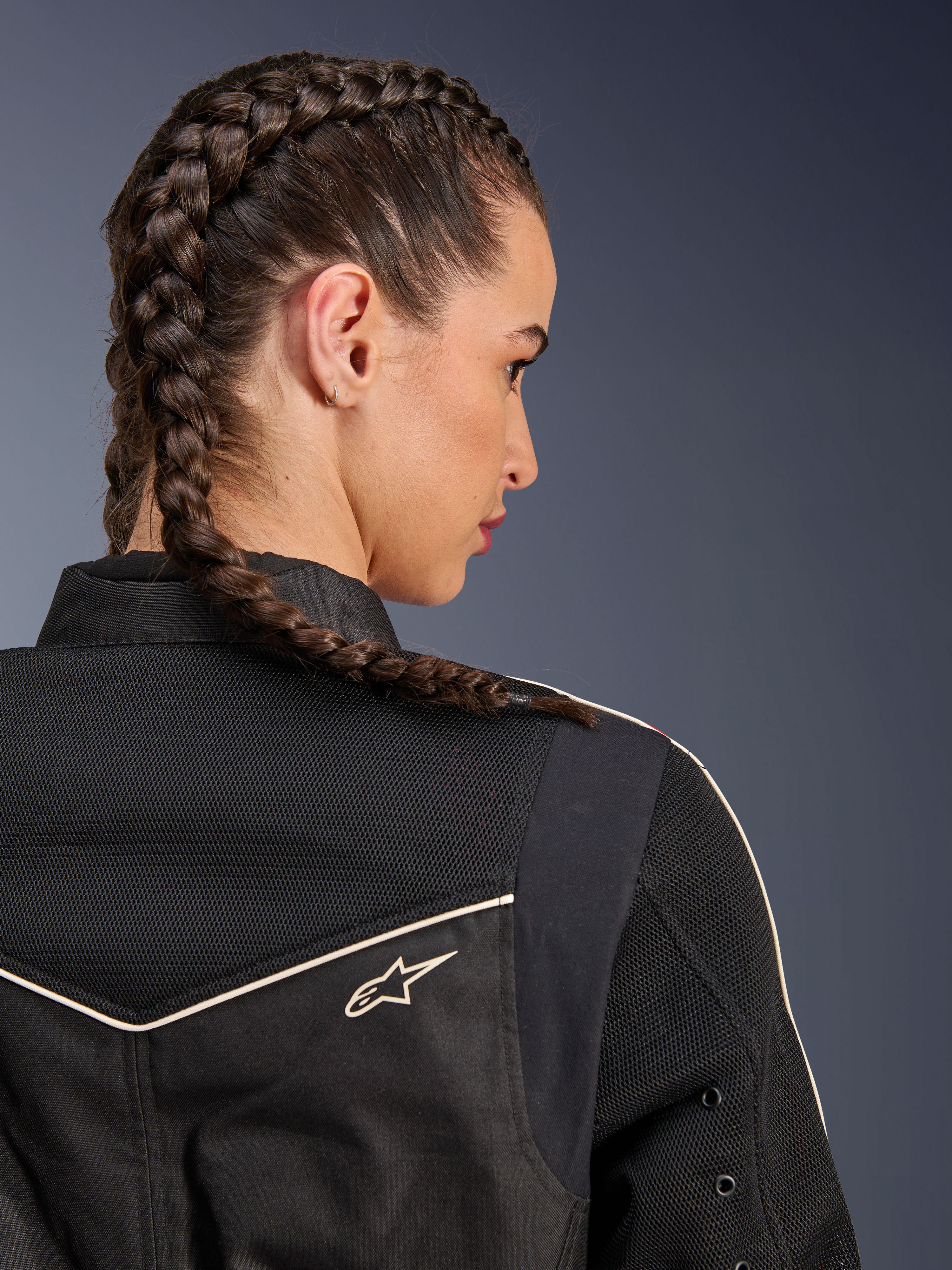 Frau in der Alpinestars Stella T-Dyno Air Jacke, Nahaufnahme der Rückansicht mit Fokus auf Schultern und Passe, Schwarz mit atmungsaktiven 3D-Mesh-Einsätzen, weißer Paspelierung und kleinem Astars-Logo, Modell mit geflochtenem Haar, urbane Motorradbekleidung