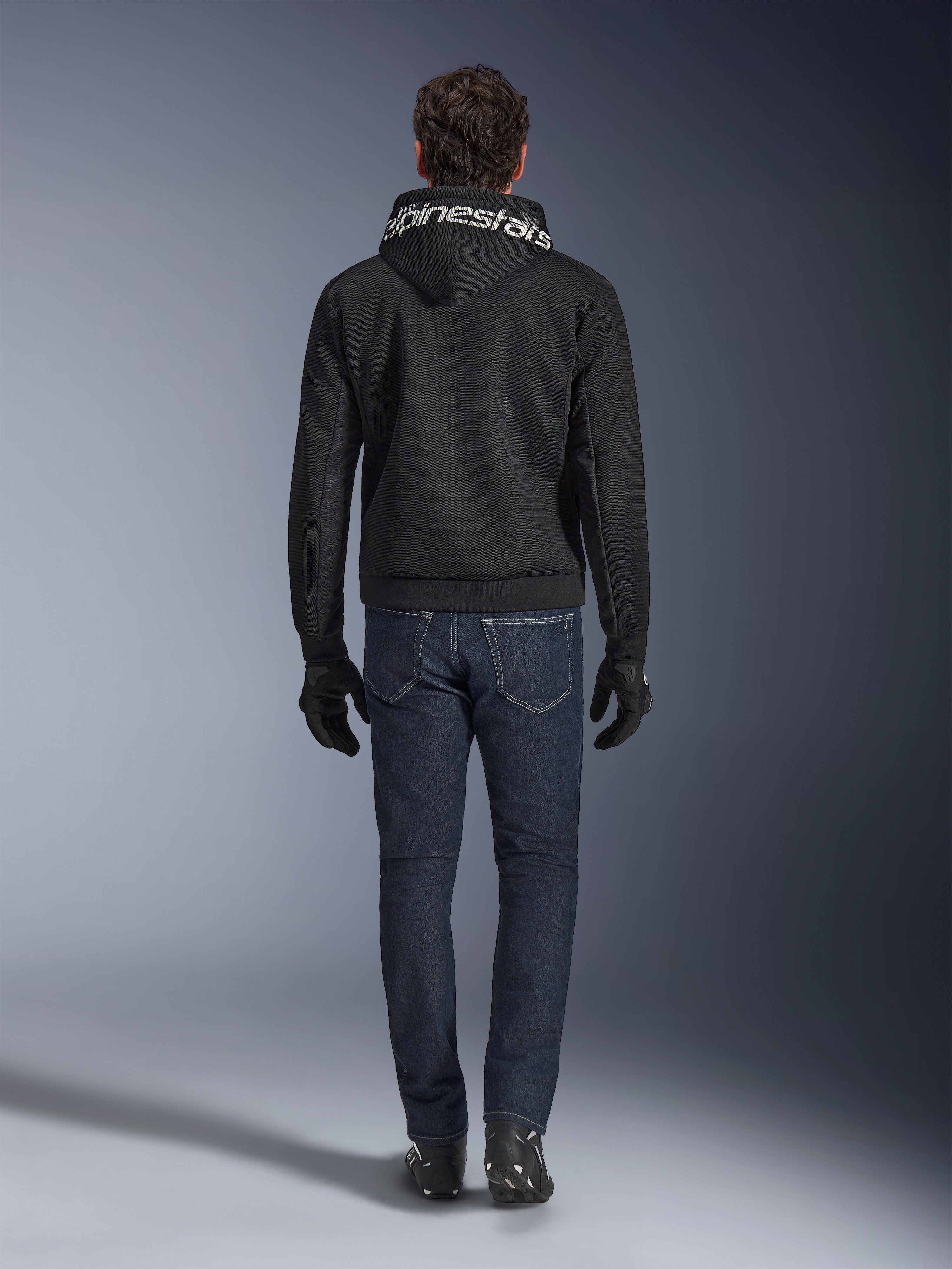 Alpinestars Chrome Superairflow Hoodie, sweat à capuche de moto noir avec logo blanc sur la capuche, vue de dos, porté avec un jean bleu foncé, des gants noirs et des bottes noires et blanches, équipement de conduite sportive urbaine