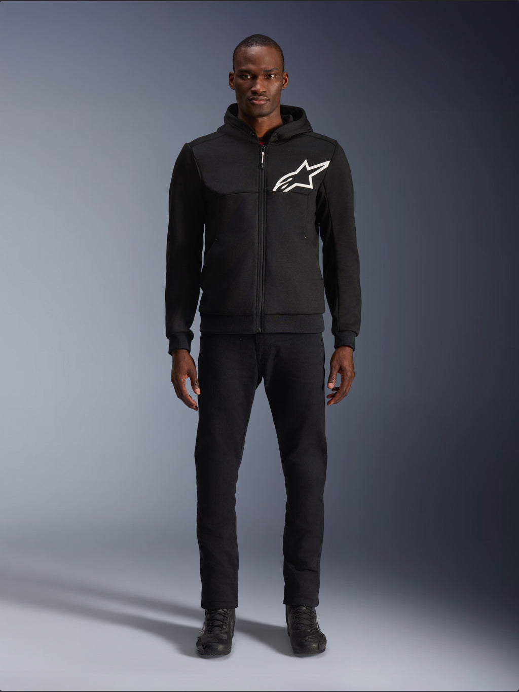 Alpinestars Chrome V2 Sport Hoodie, sweat à capuche de moto noir et blanc, avec un grand logo Alpinestars blanc sur la poitrine, fermeture éclair frontale et capuche, porté par un mannequin debout avec un pantalon et des bottes noirs, équipement de protection de style urbain.
