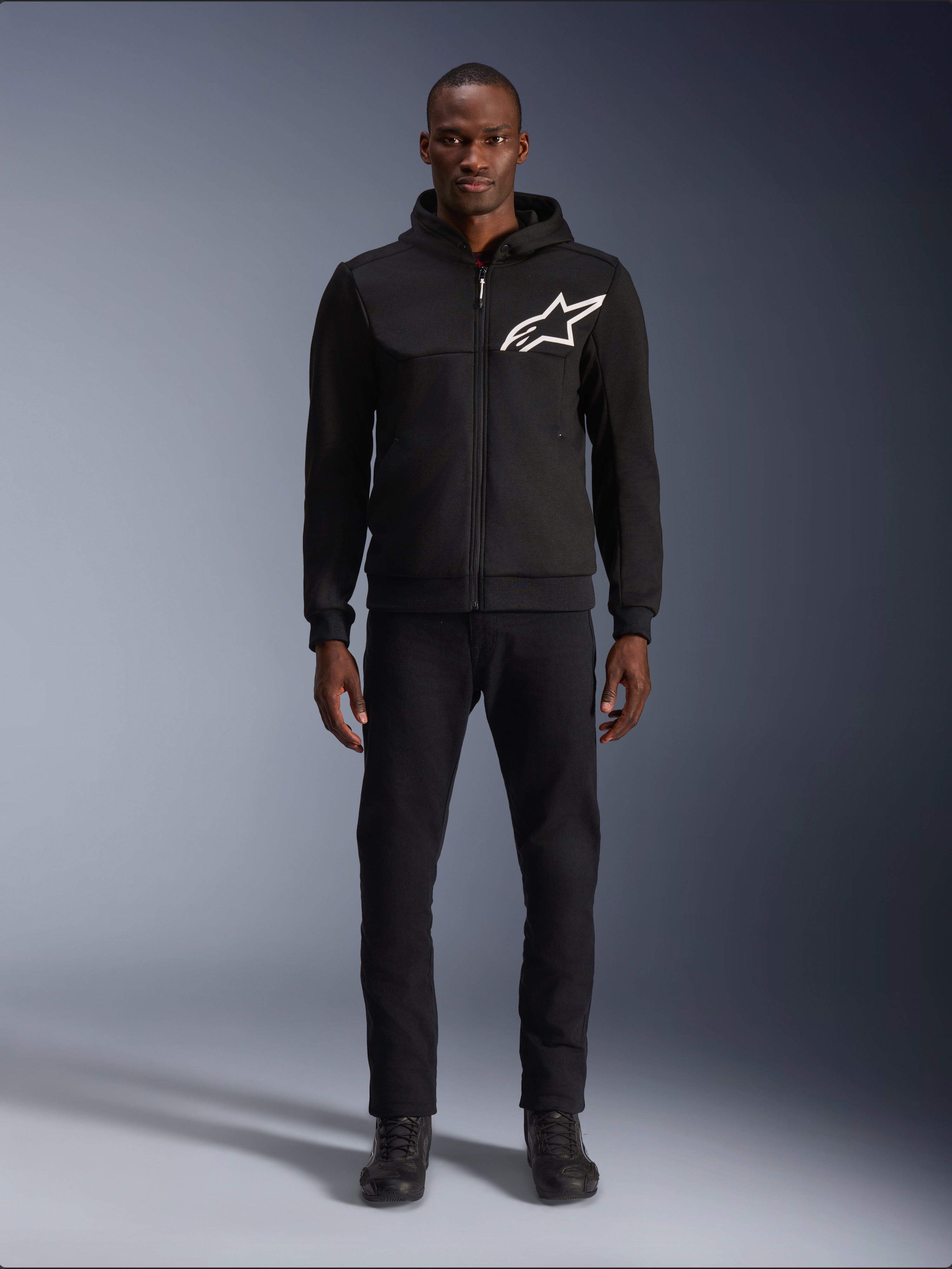 Alpinestars Chrome V2 Sport Hoodie, sweat à capuche de moto noir et blanc, avec un grand logo Alpinestars blanc sur la poitrine, fermeture éclair frontale et capuche, porté par un mannequin debout avec un pantalon et des bottes noirs, équipement de protection de style urbain.