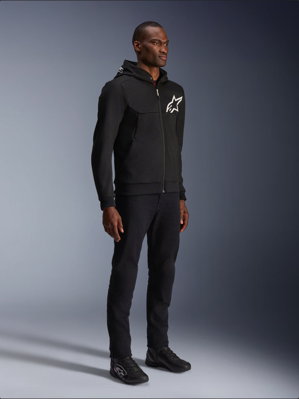 Alpinestars Chrome V2 Sport Hoodie, sweat à capuche de moto noir avec logo étoile blanc, porté par un mannequin homme avec un pantalon noir et des baskets de moto assortis, équipement de conduite au style urbain