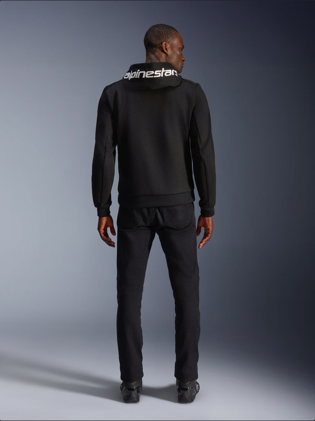 Sweat à capuche Alpinestars Chrome V2 Sport, hoodie de moto, noir, vue arrière d'un mannequin portant un sweat en polaire technique noire avec le logo Alpinestars blanc sur la capuche, associé à un pantalon et des bottes noirs, style urbain décontracté