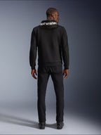 Sweat à capuche Alpinestars Chrome V2 Sport, hoodie de moto, noir, vue arrière d'un mannequin portant un sweat en polaire technique noire avec le logo Alpinestars blanc sur la capuche, associé à un pantalon et des bottes noirs, style urbain décontracté