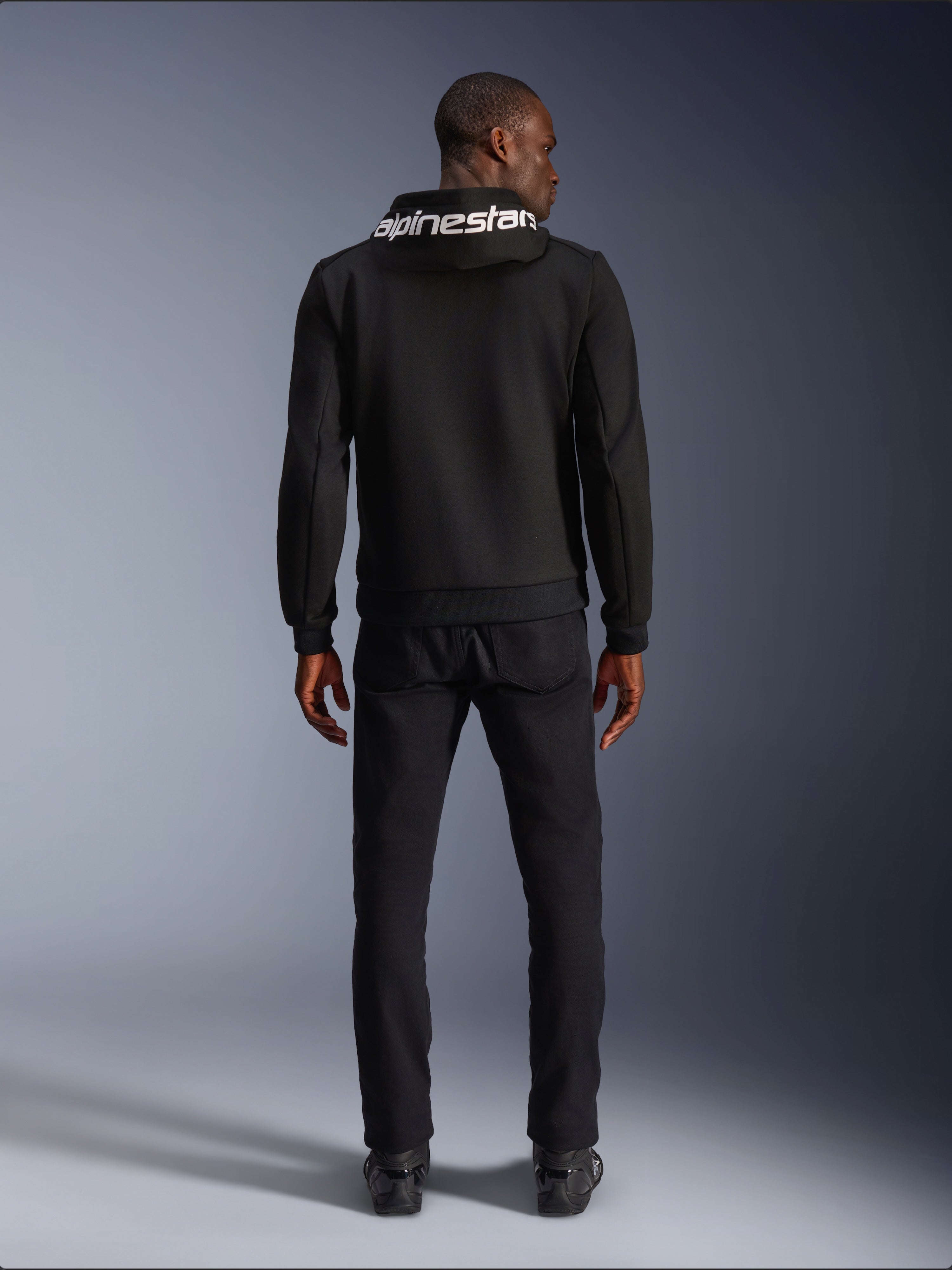 Sweat à capuche Alpinestars Chrome V2 Sport, hoodie de moto, noir, vue arrière d'un mannequin portant un sweat en polaire technique noire avec le logo Alpinestars blanc sur la capuche, associé à un pantalon et des bottes noirs, style urbain décontracté