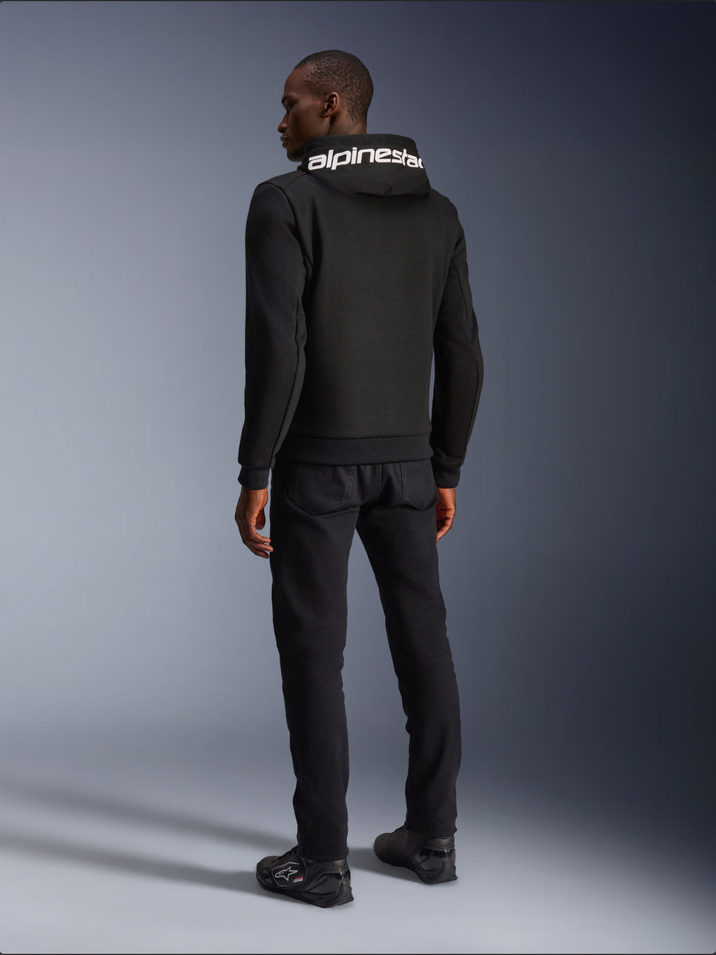 Alpinestars Chrome V2 Sport Hoodie, noir, vue de dos montrant le marquage Alpinestars blanc sur la capuche, porté avec un pantalon noir et des chaussures de moto, vêtement de moto urbain décontracté à la silhouette athlétique