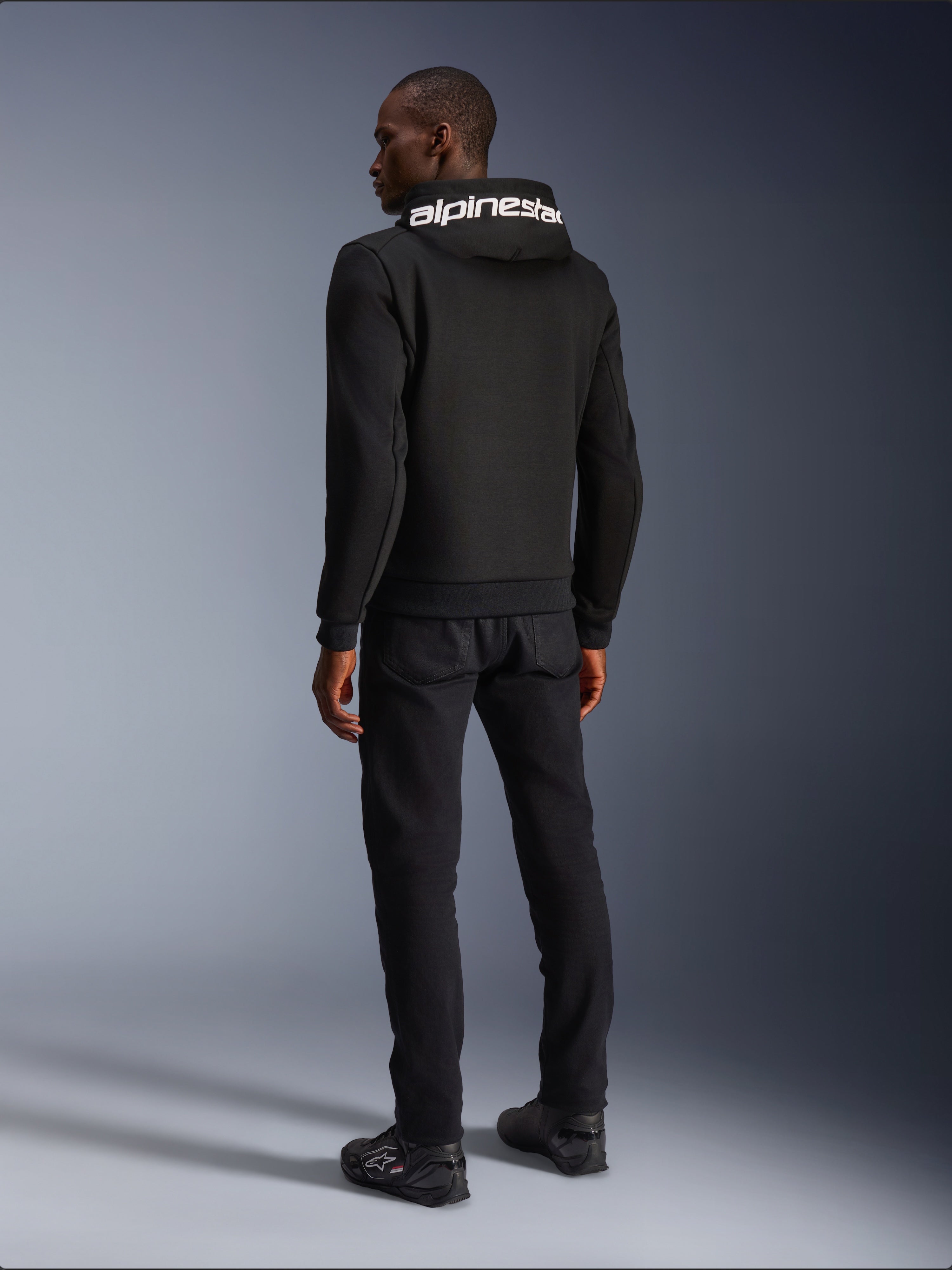 Alpinestars Chrome V2 Sport Hoodie, noir, vue de dos montrant le marquage Alpinestars blanc sur la capuche, porté avec un pantalon noir et des chaussures de moto, vêtement de moto urbain décontracté à la silhouette athlétique