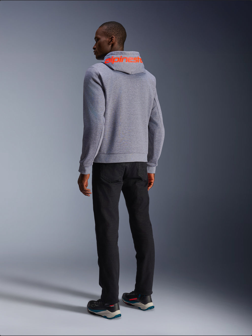 Alpinestars Chrome V2 Sport Hoodie in Gray Heather, Rückansicht mit großem Alpinestars-Schriftzug in Red Fluo auf der Kapuze, getragen von einem Model mit schwarzer Hose und dunklen Motorradschuhen, lässige urbane Motorradbekleidung