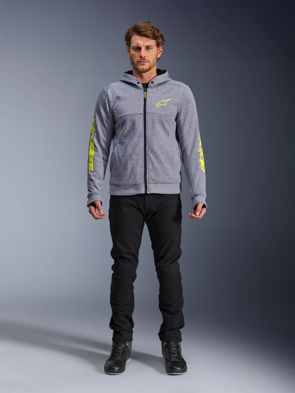 Sweat à capuche Alpinestars Chrome V2 Sport, gris chiné avec accents jaune fluo, vêtement de moto urbain, présenté avec un pantalon noir et des chaussures de moto noires, polaire technique avec marquage jaune vif sur les manches et la poitrine, style urbain décontracté pour la conduite