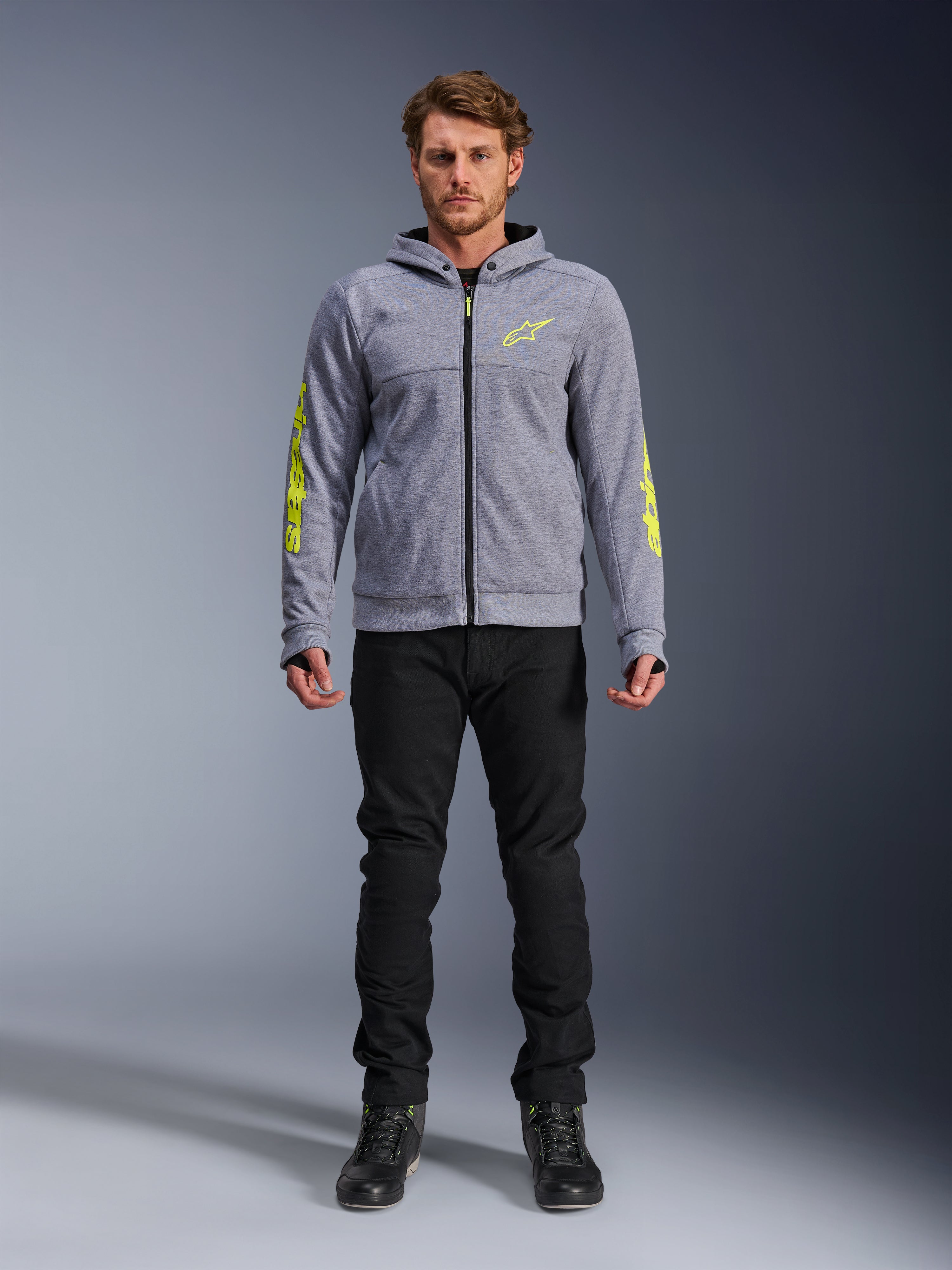 Sweat à capuche Alpinestars Chrome V2 Sport, gris chiné avec accents jaune fluo, vêtement de moto urbain, présenté avec un pantalon noir et des chaussures de moto noires, polaire technique avec marquage jaune vif sur les manches et la poitrine, style urbain décontracté pour la conduite