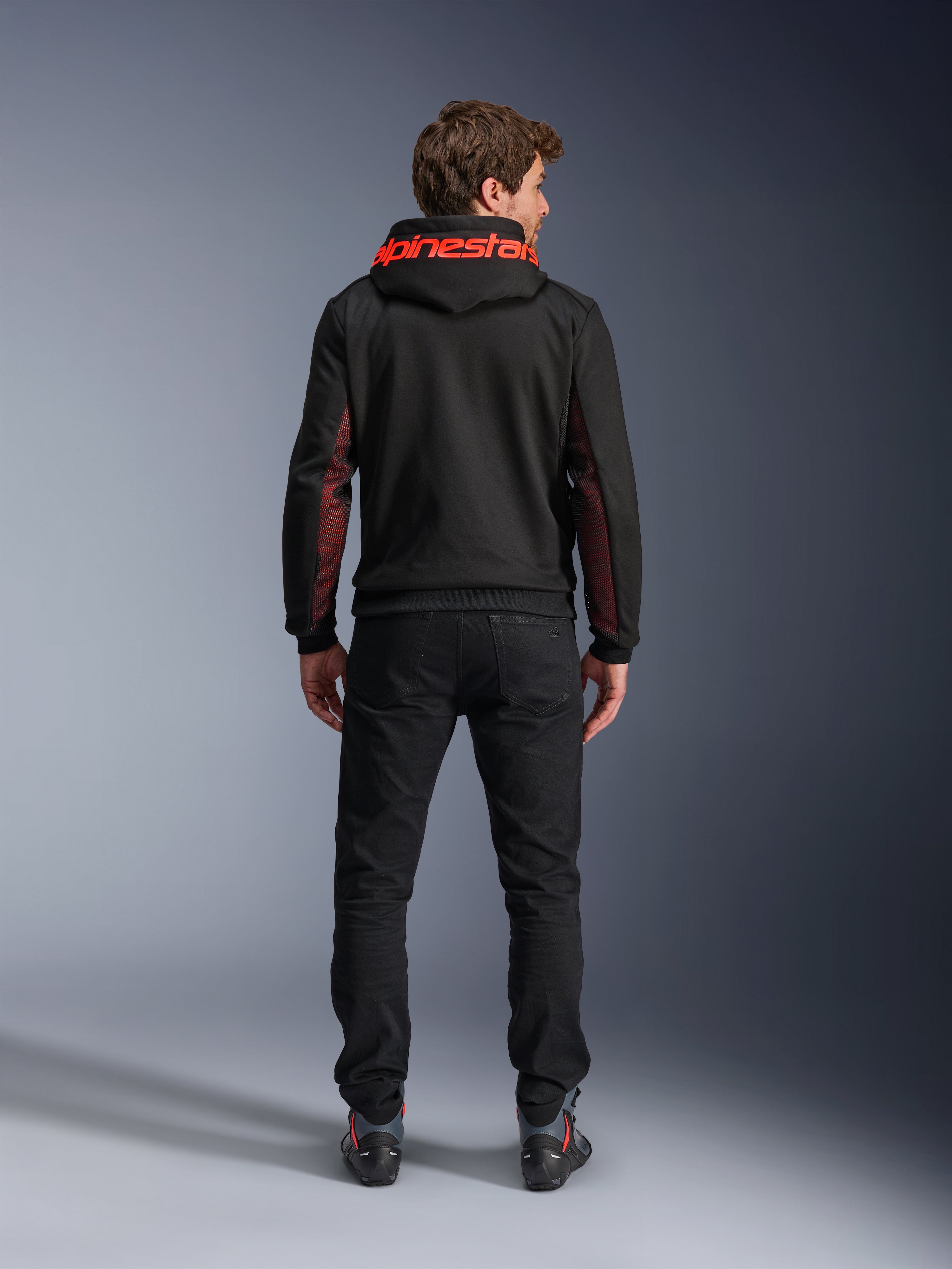 Alpinestars Chrome Air Sport Hoodie, Schwarz und Fluorot, Rückansicht eines Mannes, der eine lässige Motorradjacke trägt, mit rotem Alpinestars-Branding auf der Kapuze und roten Mesh-Einsätzen an den Ärmeln, kombiniert mit einer schwarzen Hose.