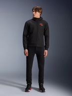 Alpinestars MM93 Radium Tech Hoodie, schwarzer urbaner Motorrad-Hoodie, mit rotem 93-Logo und weißem Stern-Logo auf der Brust, getragen von einem Model mit schwarzer Hose und passenden Motorradschuhen, elegante winddichte technische Shell für das urbane Fahren.
