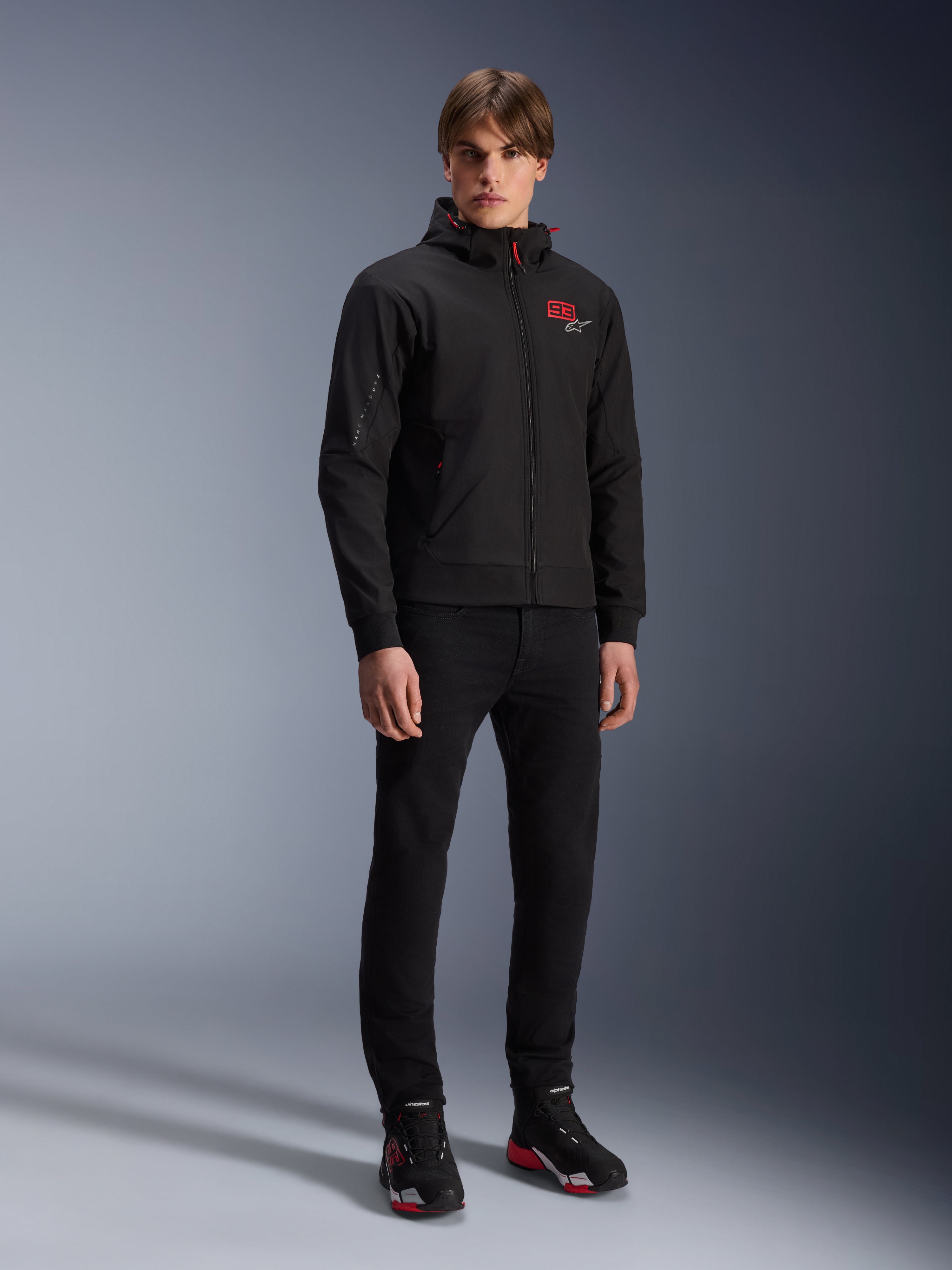 Alpinestars MM93 Radium Tech Hoodie, urbane Motorradjacke, Schwarz mit leuchtend roten Akzenten, getragen von einem Model mit schwarzer Hose und Motorradschuhen, mit rotem „93“-Logo auf der Brust und Marc Marquez Branding am Ärmel, lässige technische Streetwear für die Stadt