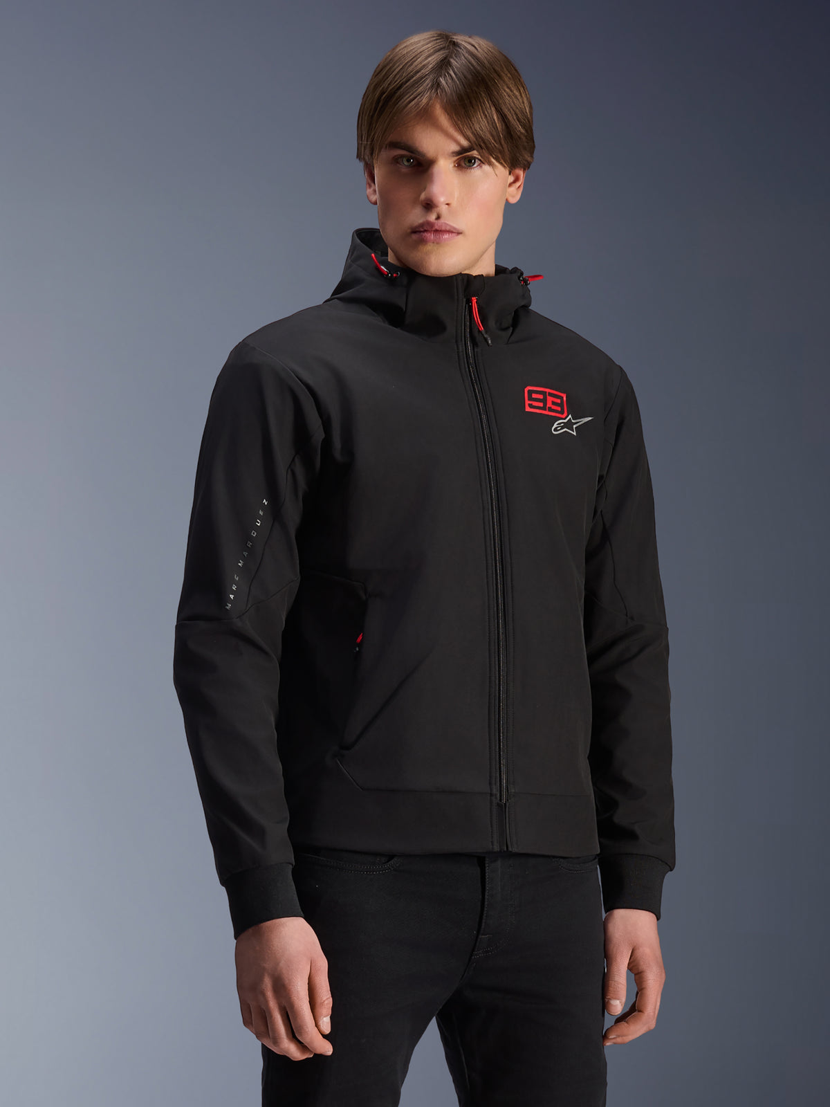 MM93 Radium Tech Hoodie, zwarte urban motorjas met ritssluiting, rood 93-nummer en wit Alpinestars-logo op de borst, gedragen door model met zwarte broek, voorzien van een afneembare capuchon met rode trekkoorden en branding op de mouw, casual motorsport-geïnspireerd ontwerp