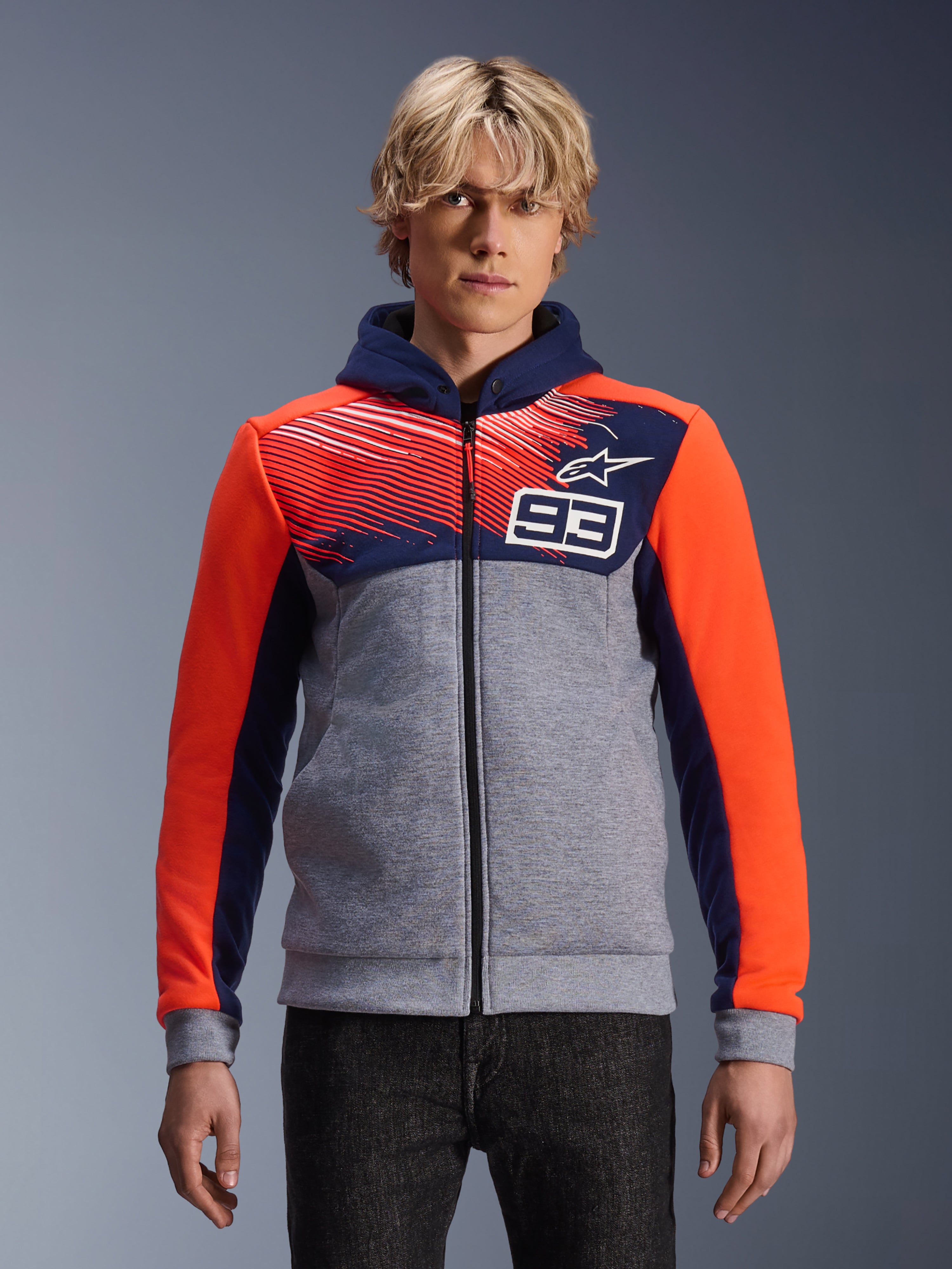 Alpinestars MM93 Chrome V2 Sport Hoodie, motorhoodie met rits, grijs gemêleerd met donkerblauwe borst en capuchon, feloranje mouwen, nummer 93 op de borst met gestreept patroon en sterlogo, gedragen door model met donkere denim, Marc Marquez-collectie.