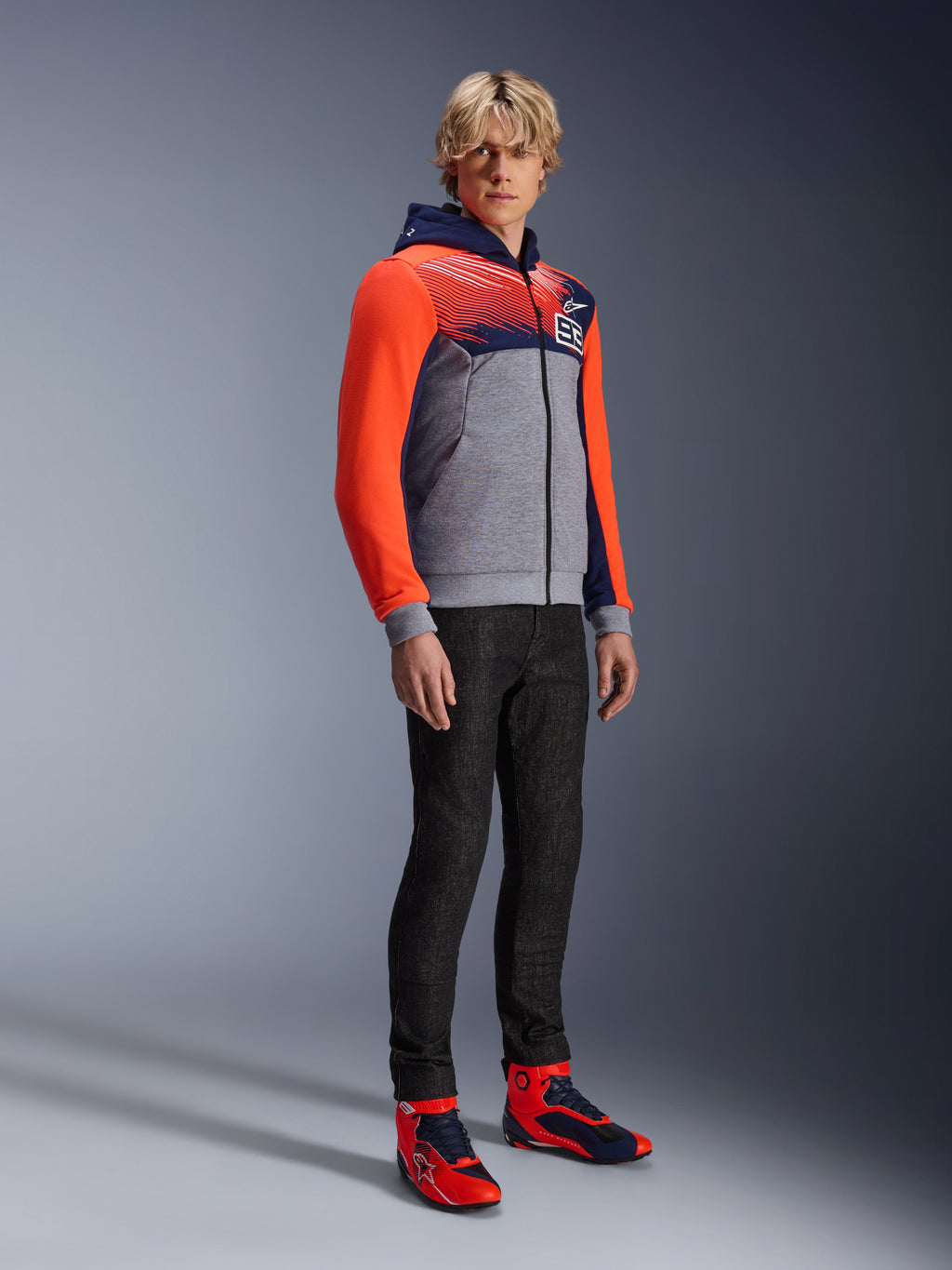 Sweat à capuche sport Alpinestars MM93 Chrome V2, sweat de moto urbain, corps gris chiné avec poitrine bleu marine et manches rouge fluo, logo 93 sur la poitrine, porté par un mannequin avec un pantalon noir ajusté et des chaussures de moto montantes coordonnées rouges et bleues, coupe athlétique moderne pour la conduite urbaine