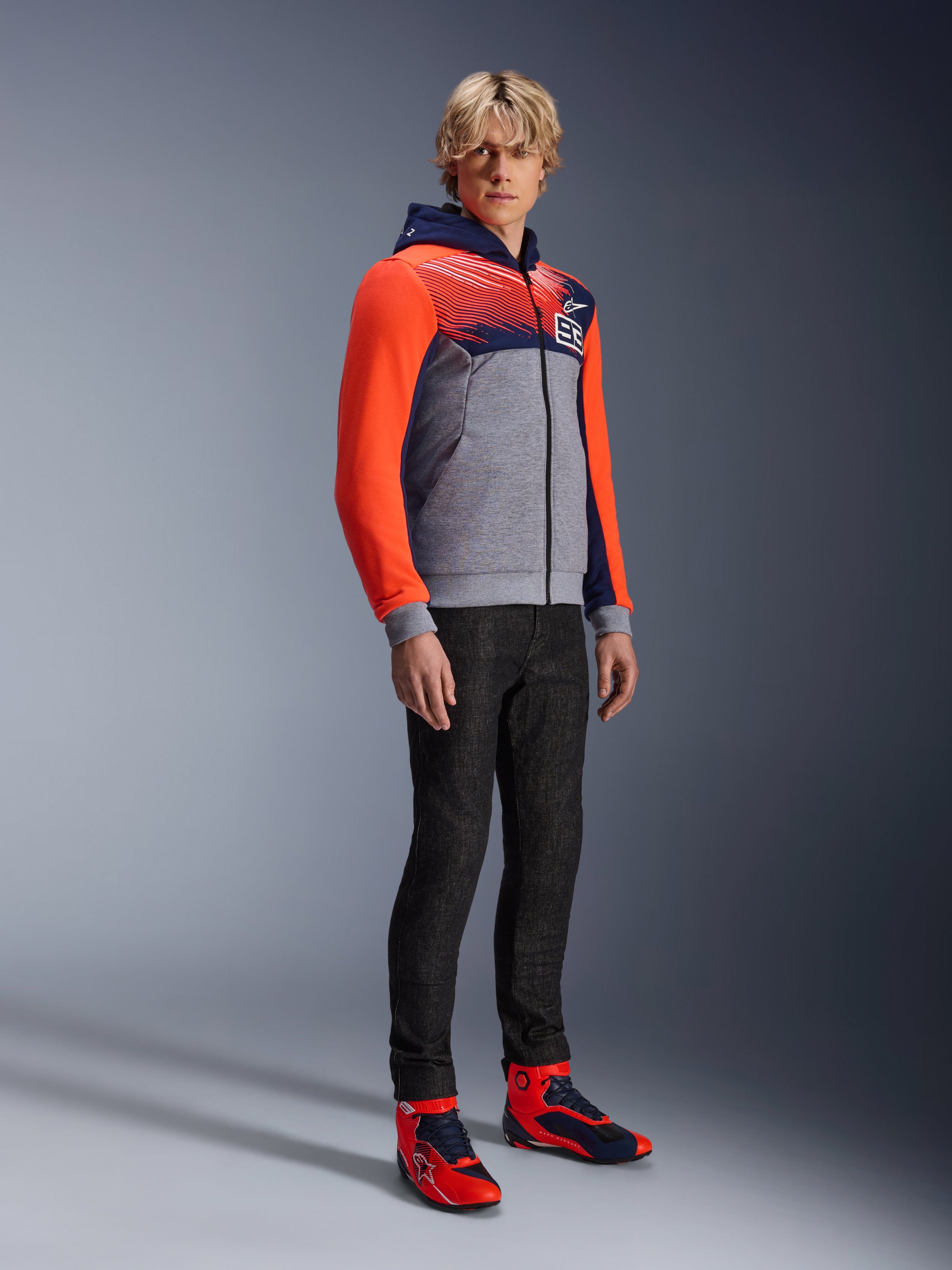 Sweat à capuche sport Alpinestars MM93 Chrome V2, sweat de moto urbain, corps gris chiné avec poitrine bleu marine et manches rouge fluo, logo 93 sur la poitrine, porté par un mannequin avec un pantalon noir ajusté et des chaussures de moto montantes coordonnées rouges et bleues, coupe athlétique moderne pour la conduite urbaine