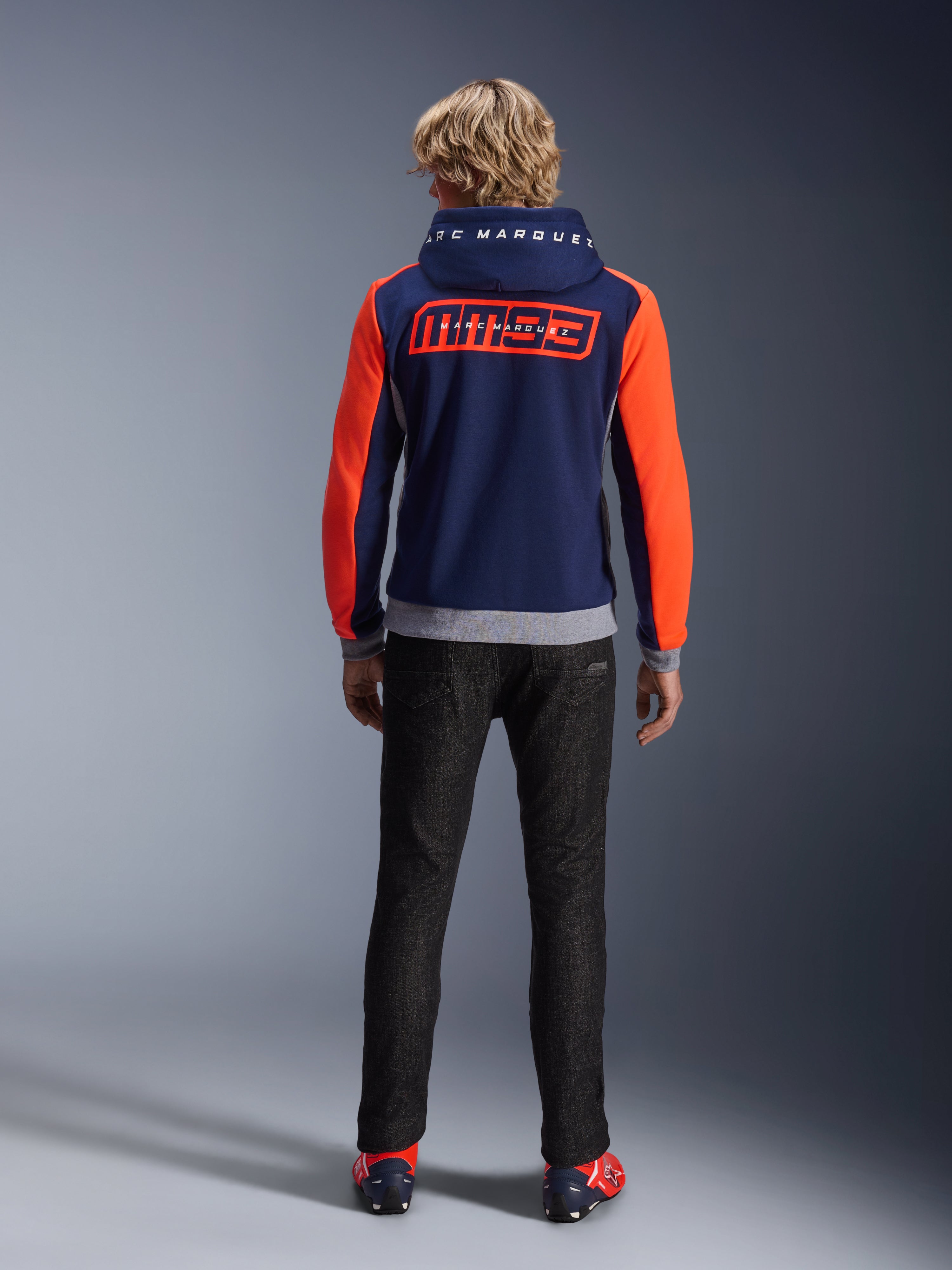 Modèle de dos portant le sweat à capuche Alpinestars MM93 Chrome V2 Sport, bleu marine avec manches rouge fluo éclatant et détails gris chiné, grands logos MM93 et Marc Marquez dans le dos et sur la capuche, sweat à capuche de moto décontracté associé à un pantalon noir