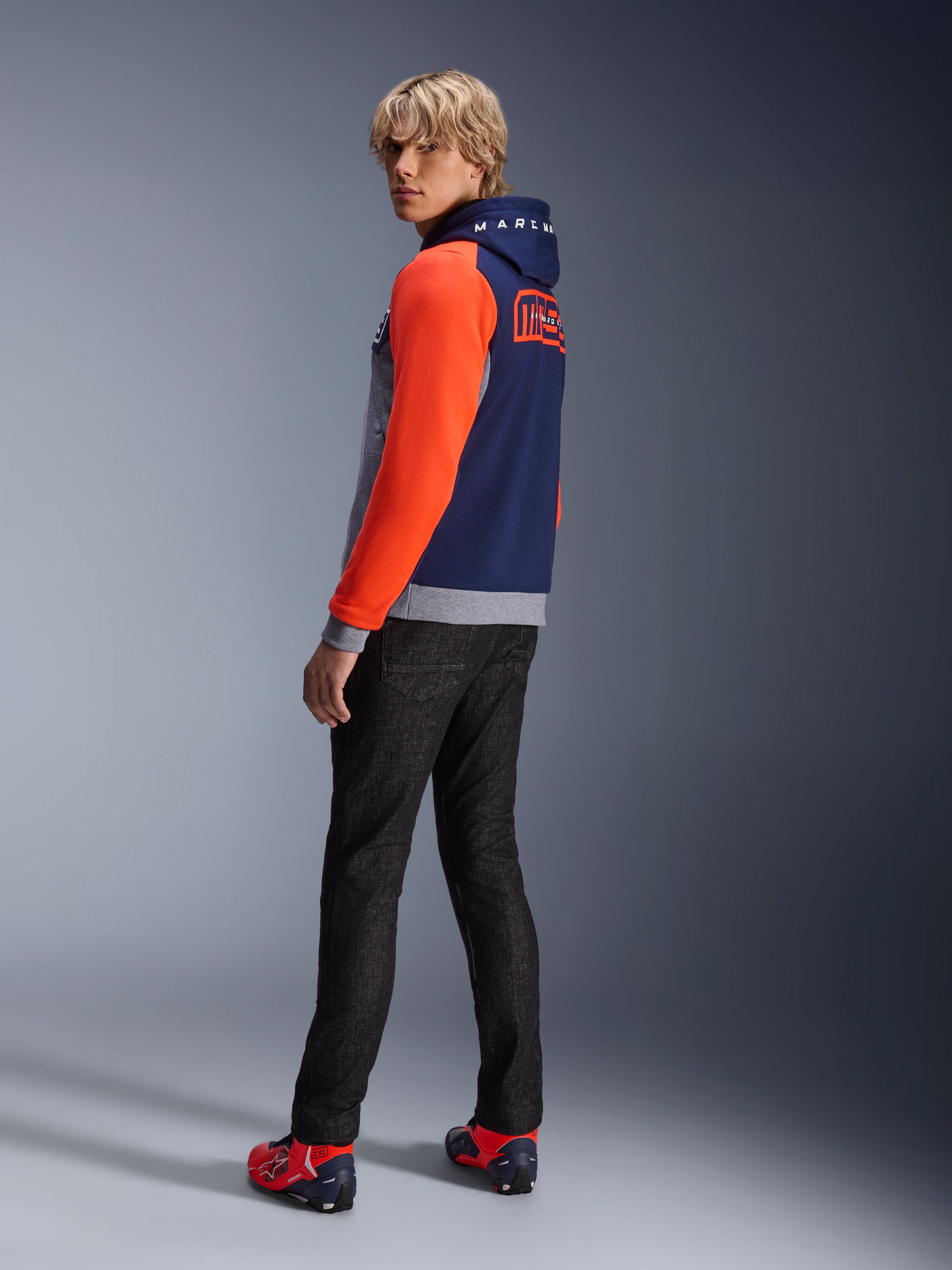 Alpinestars MM93 Chrome V2 Sport Hoodie, bleu marine, gris chiné et rouge fluo, mannequin portant le sweat à capuche avec des manches rouge vif et le logo MM93 Marc Marquez au dos, associé à un jean foncé et des chaussures de moto rouges, équipement de moto urbain décontracté