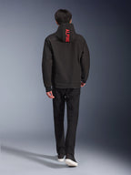 Alpinestars Radium Tech Hoodie, urbane Motorradjacke, Schwarz, Rückansicht mit leuchtend rotem „ALPINE“-Schriftzug auf der Kapuze, getragen mit schwarzer Hose und Sneakern, schlanke technische Shell für den Pendlerverkehr.