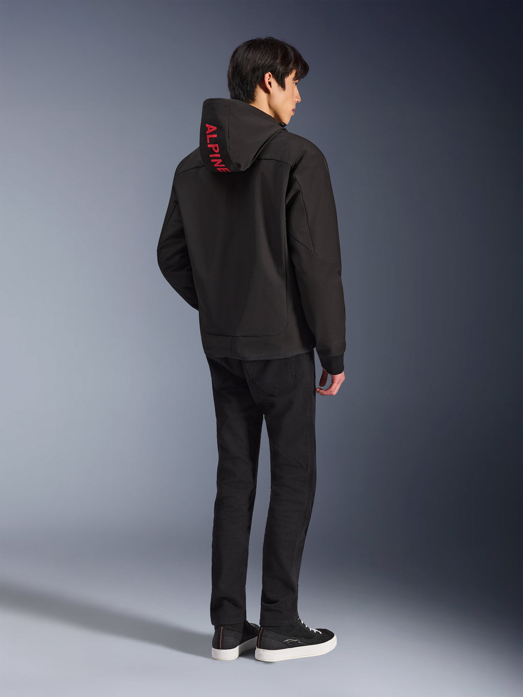 Alpinestars Radium Tech Hoodie, Schwarz mit rotem Alpinestars-Branding auf der Kapuze, Rückansicht an einem männlichen Model, getragen mit schwarzer Hose und Sneakern, technische urbane Fahrausrüstung