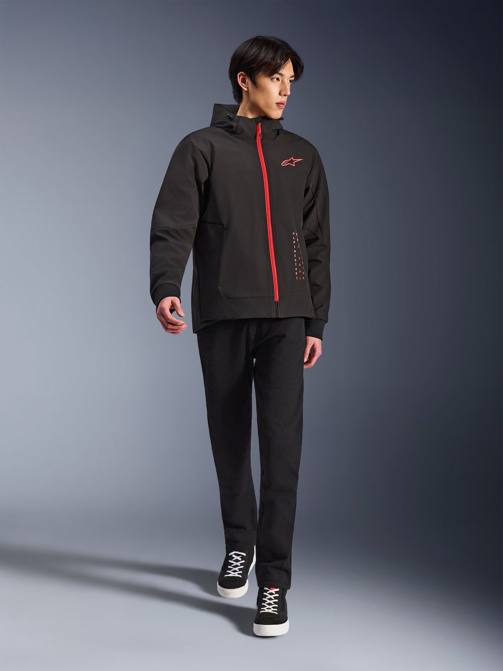 Alpinestars Radium Tech Hoodie in Black Bright Red, urbane Motorradjacke getragen von einem Model, mit rotem Frontreißverschluss und Brustlogo, kombiniert mit schwarzer Hose und Sneakern für einen modernen Pendler-Look.