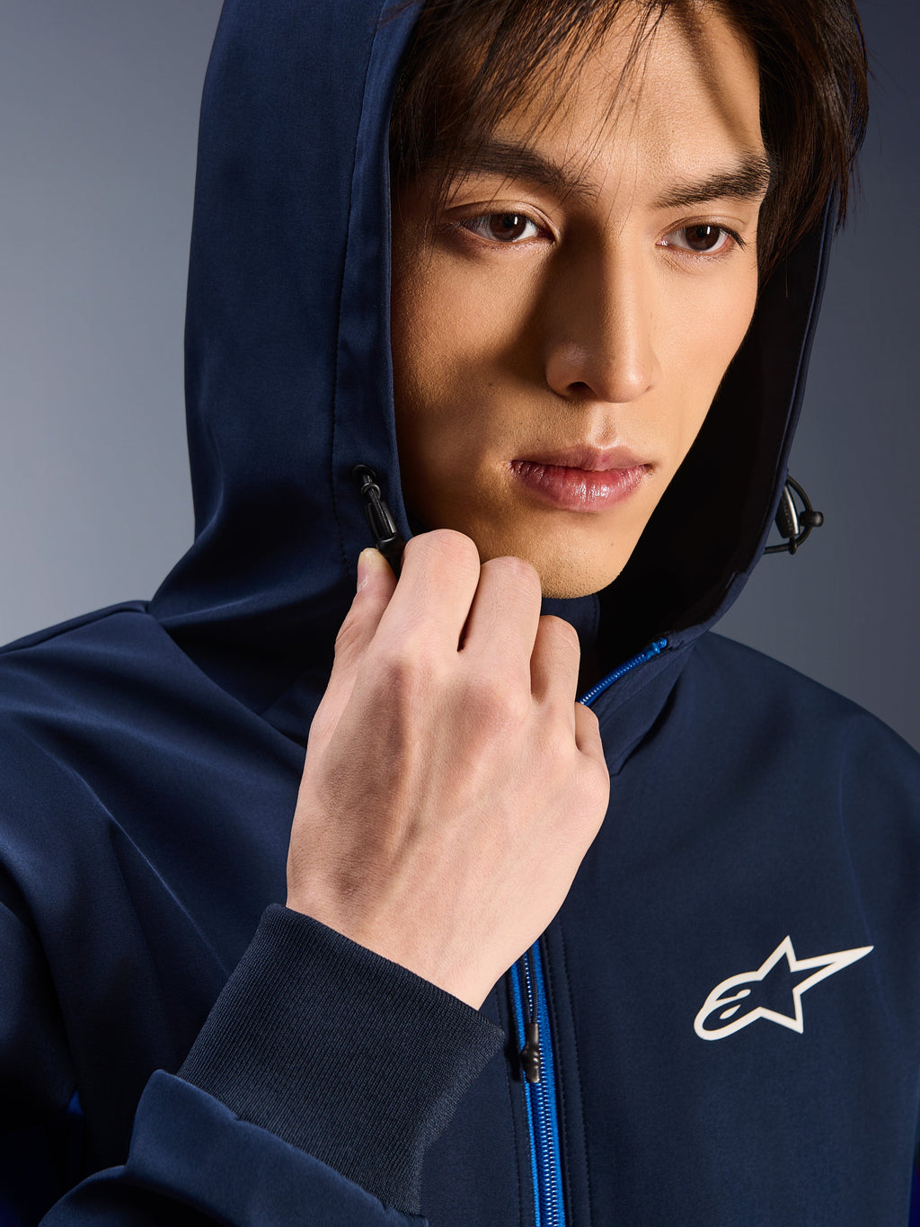 Alpinestars Radium Tech Hoodie, Night Navy Royal Blue, Nahaufnahme eines Mannes mit aufgesetzter Kapuze, weißes Astars-Logo auf der Brust, blauer Reißverschluss, urbane technische Motorradjacke
