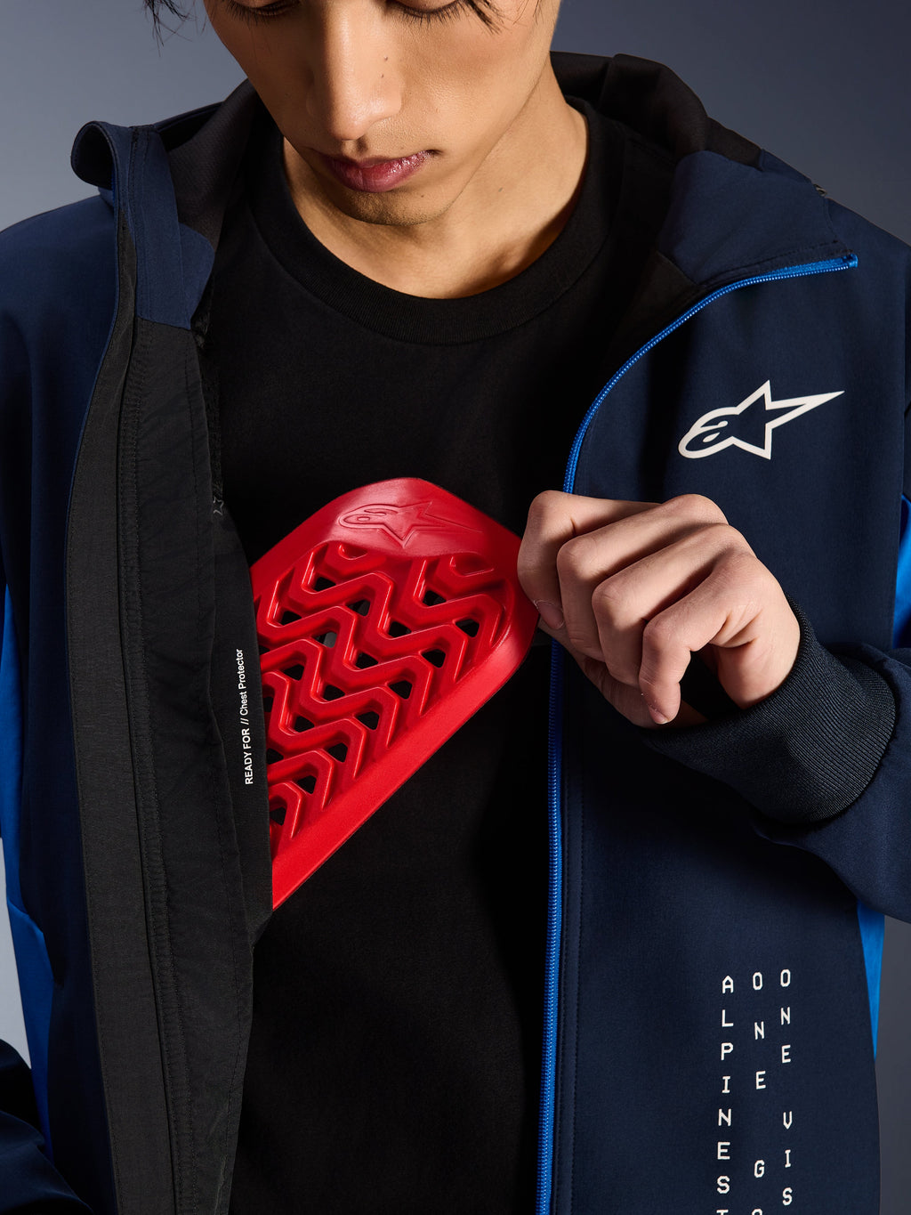 Alpinestars Radium Tech Hoodie in Night Navy und Royal Blue, Nahaufnahme des Einsetzens eines roten Nucleon Brustprotektors in die Innentasche, mit dem Schriftzug „READY FOR // Chest Protector“ auf dem Futter und einem weißen Stern-Logo auf der Brust.