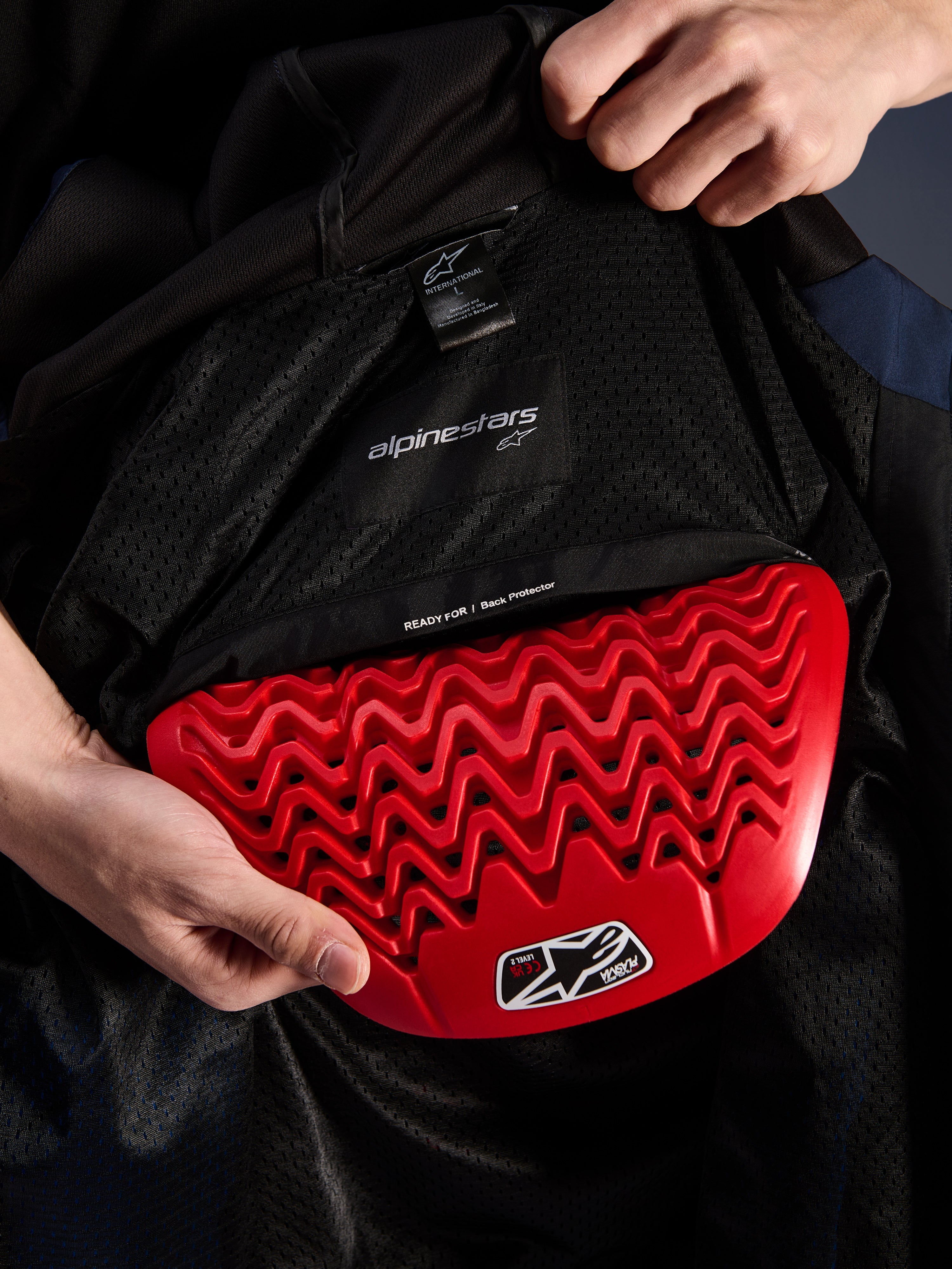 Alpinestars Radium Tech Hoodie Innendetail, roter Nucleon Plasma Rückenprotektor wird in die Tasche eingesetzt, schwarzes Mesh-Futter mit 'READY FOR | Back Protector' Text, wellenförmiges Waben-Design, technische Schutzmerkmale einer urbanen Motorradjacke