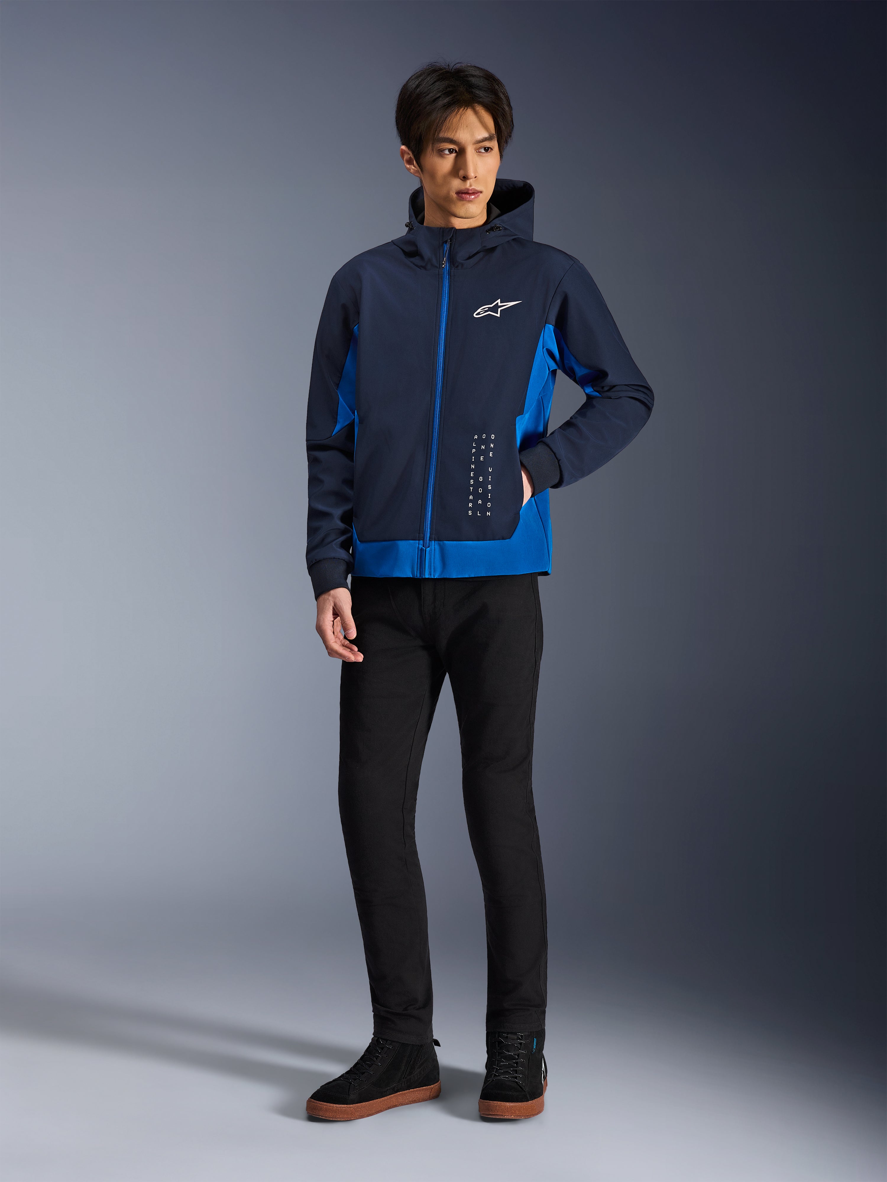 Alpinestars Radium Tech Hoodie, urbane Motorradjacke, Night Navy und Royal Blue, getragen mit schwarzer Hose und schwarzen Sneakern, mit weißem Alpinestars-Logo auf der Brust und vertikalen Textdetails, lässige sportliche Passform für Stadtfahrten