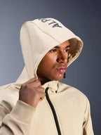 Alpinestars Radium Tech Hoodie in Mastic Aluminum, Nahaufnahme eines Mannes mit aufgesetzter Kapuze, Alpinestars-Branding auf der Oberseite, urbane Motorradjacke mit technischer winddichter Shell, moderne Streetwear-Ästhetik für Stadtfahrten
