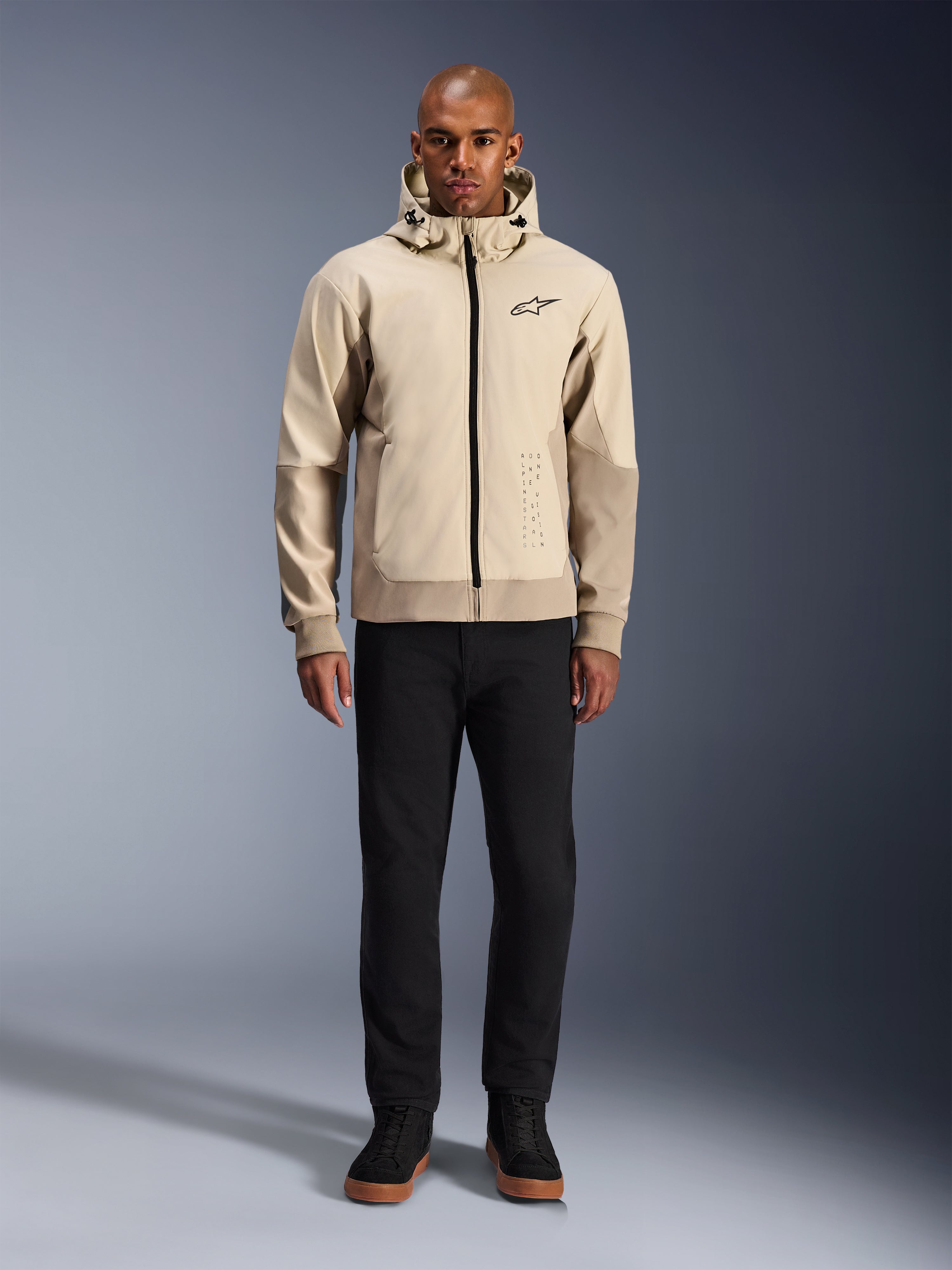 Alpinestars Radium Tech Hoodie in Mastic Aluminum, urbaner Motorrad-Hoodie, getragen von einem männlichen Model mit schwarzer Hose und schwarzen Stiefeln, mit durchgehendem schwarzem Reißverschluss, kleinem Brustlogo und athletischer Passform für Stadtfahrten.