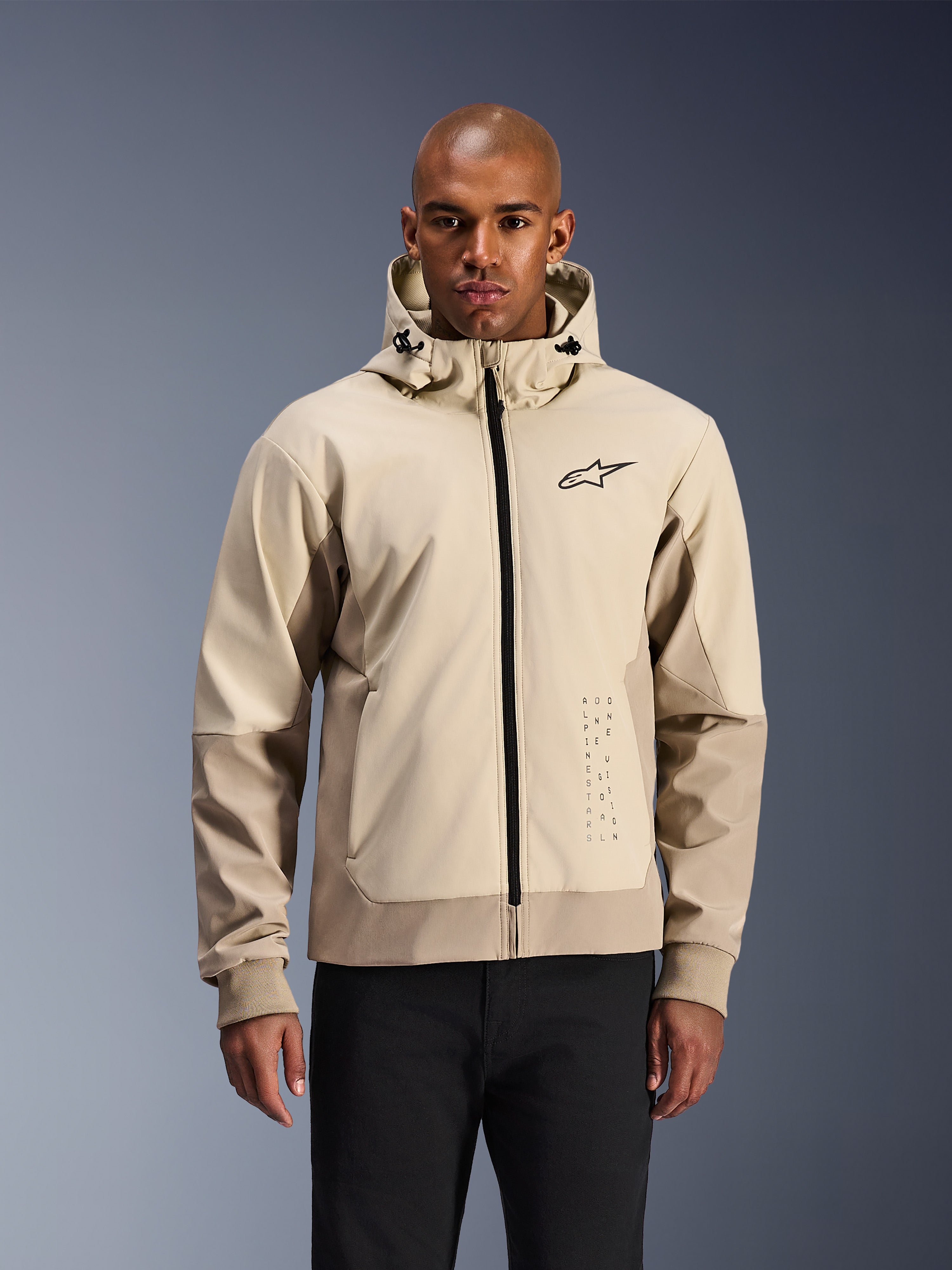 Alpinestars Radium Tech Hoodie in Mastic Aluminum, hellbeige und hellbraune urbane Motorradjacke, schwarzer Frontreißverschluss und kleines Brustlogo, präsentiert von einem Mann in schwarzer Hose, technisches winddichtes Außenmaterial mit modernem Streetwear-Styling