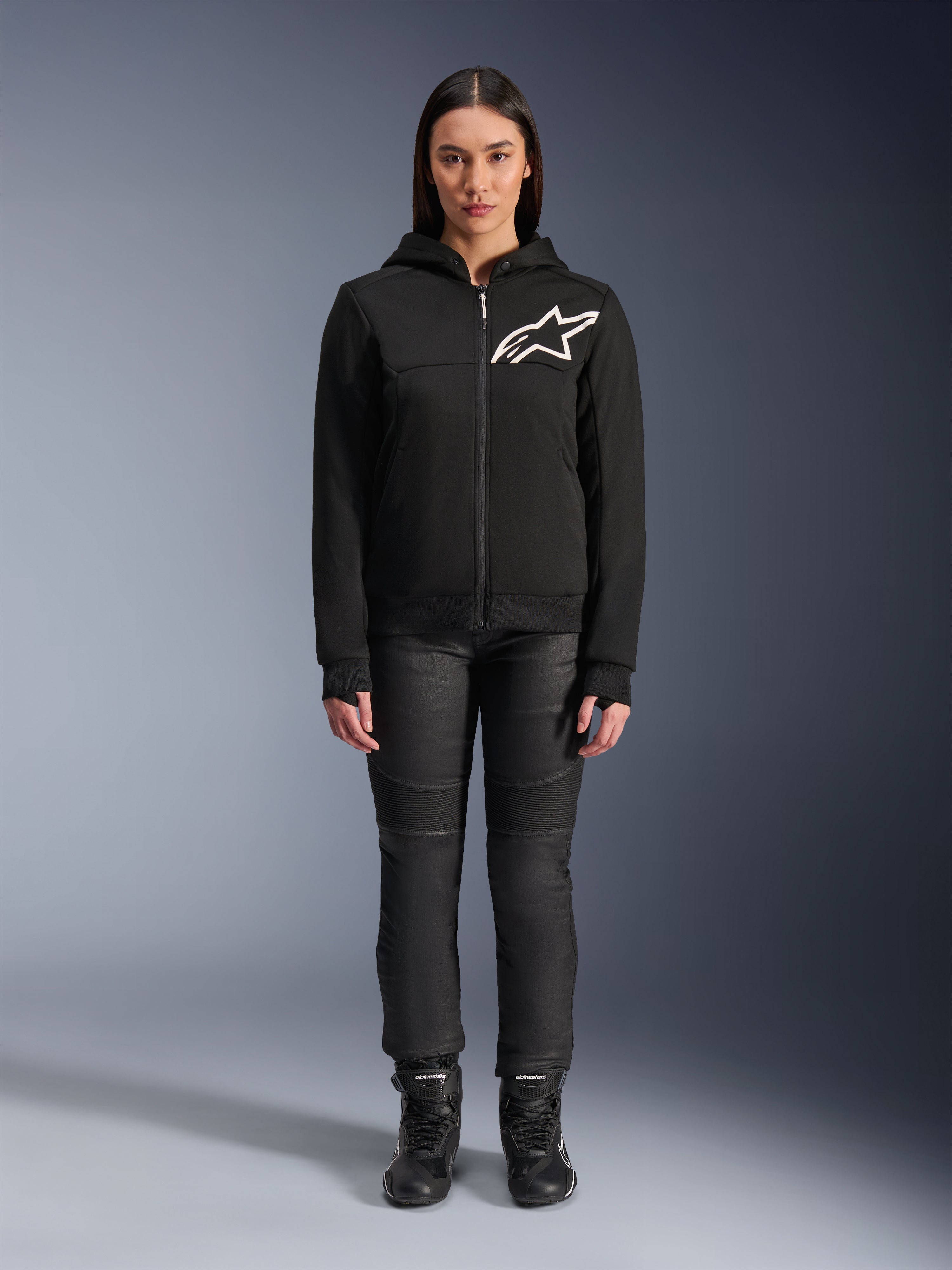 Femme portant le sweat à capuche Alpinestars Stella Chrome V2 Sport, noir avec logo Astars blanc sur la poitrine, associé à un pantalon de moto technique et des bottes noirs, design de vêtement de moto urbain.