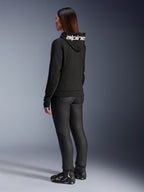 Femme portant le sweat à capuche Alpinestars Stella Chrome V2 Sport, vue de dos, sweat à capuche de moto noir avec logo Alpinestars blanc sur la capuche, assorti à un pantalon de moto technique noir et des chaussures de moto noires, tenue urbaine décontractée