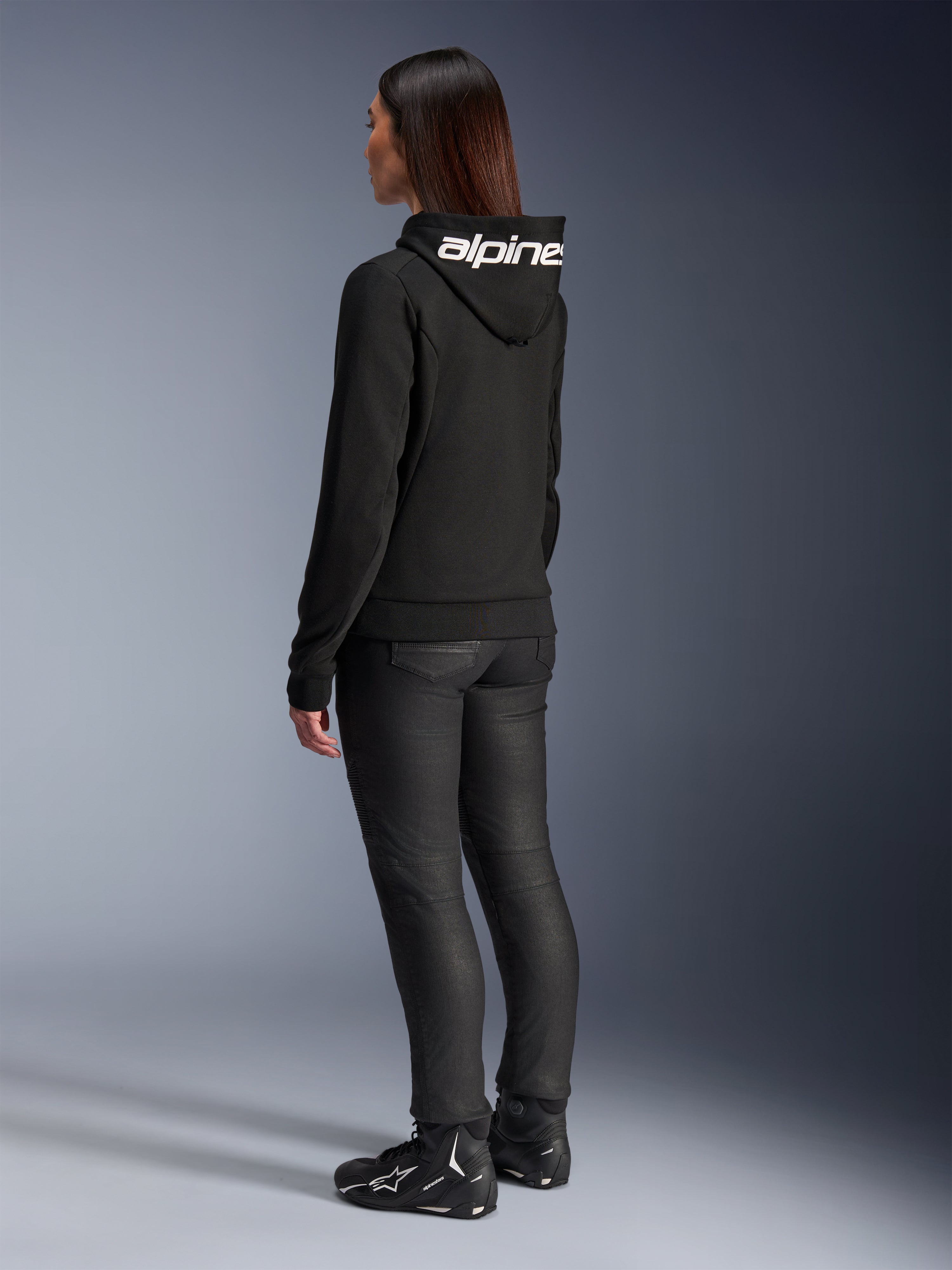 Femme portant le sweat à capuche Alpinestars Stella Chrome V2 Sport, vue de dos, sweat à capuche de moto noir avec logo Alpinestars blanc sur la capuche, assorti à un pantalon de moto technique noir et des chaussures de moto noires, tenue urbaine décontractée