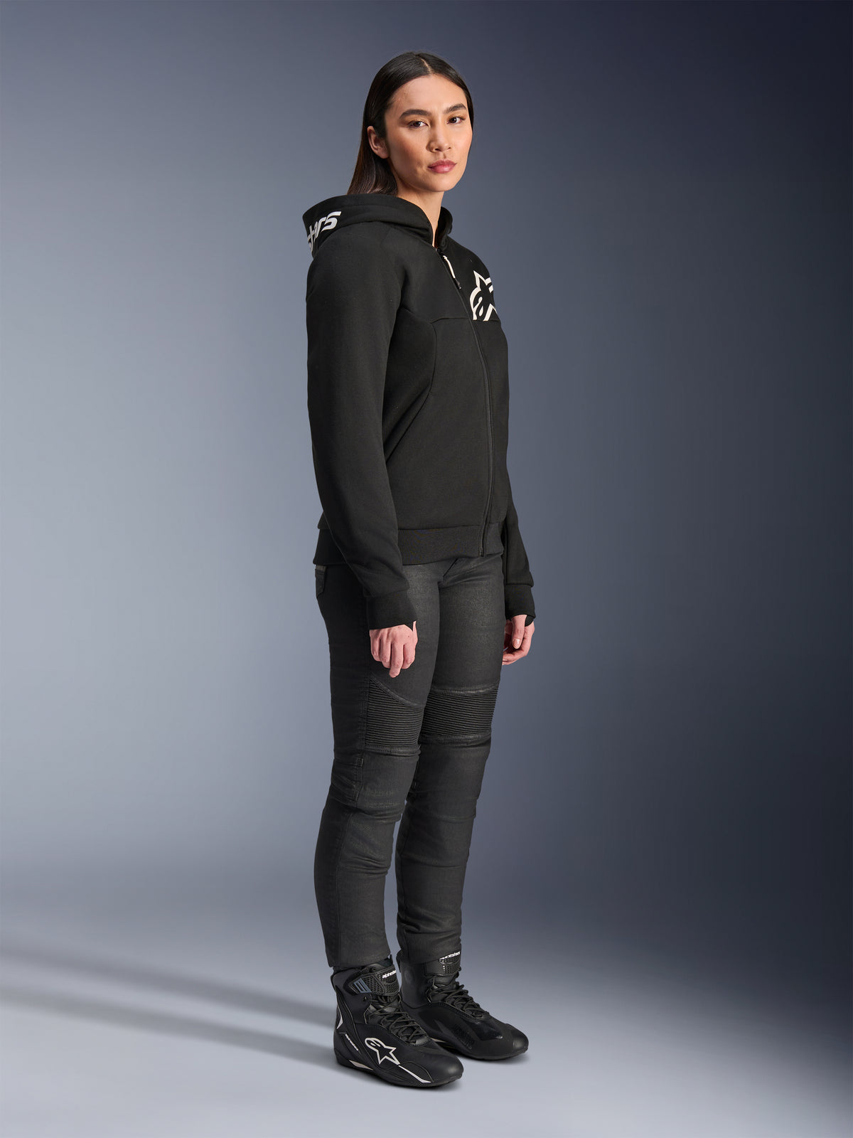 Femme portant le sweat à capuche Alpinestars Stella Chrome V2 Sport noir avec logos blancs, associé à un pantalon de moto et des bottes noirs, équipement de moto urbain pour femme à la coupe athlétique décontractée