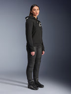 Femme portant le sweat à capuche Alpinestars Stella Chrome V2 Sport noir avec logos blancs, associé à un pantalon de moto et des bottes noirs, équipement de moto urbain pour femme à la coupe athlétique décontractée