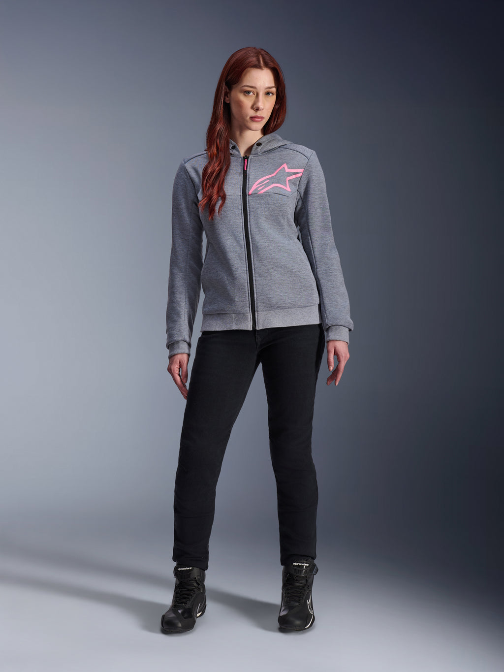 Alpinestars Stella Chrome V2 Sport Hoodie, Gray Heather Pink Fluo, gedragen door een vrouw met zwarte broek en laarzen, met een roze Alpinestars-sterlogo op de borst, urban motorkleding met volledige ritssluiting, lichtgewicht tech fleece met een casual atletische pasvorm.