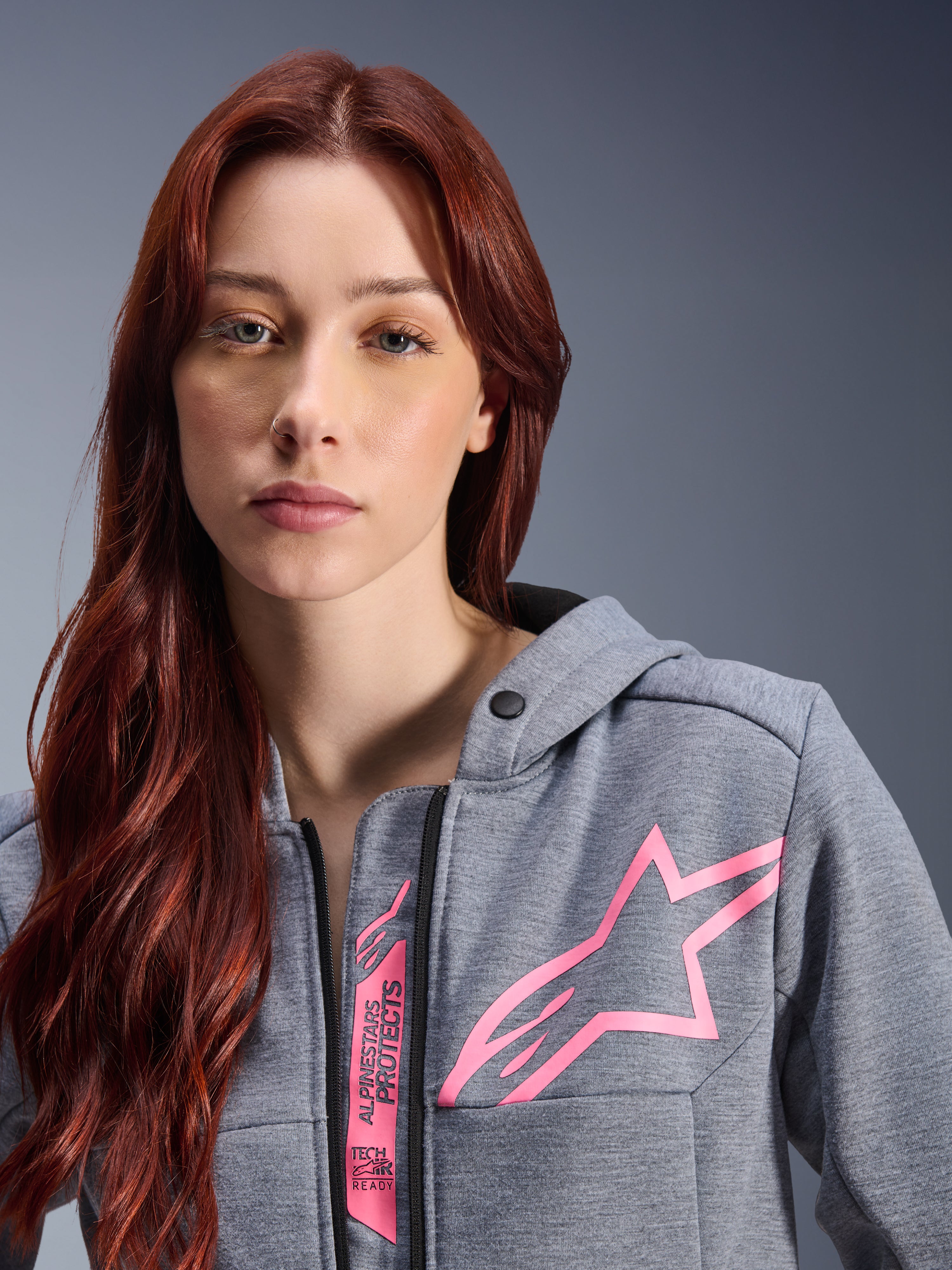 Vrouw draagt Alpinestars Stella Chrome V2 Sport Hoodie, Gray Heather Pink Fluo, close-up opname van het roze Alpinestars sterlogo en Tech-Air Ready branding, stedelijke motorkleding.