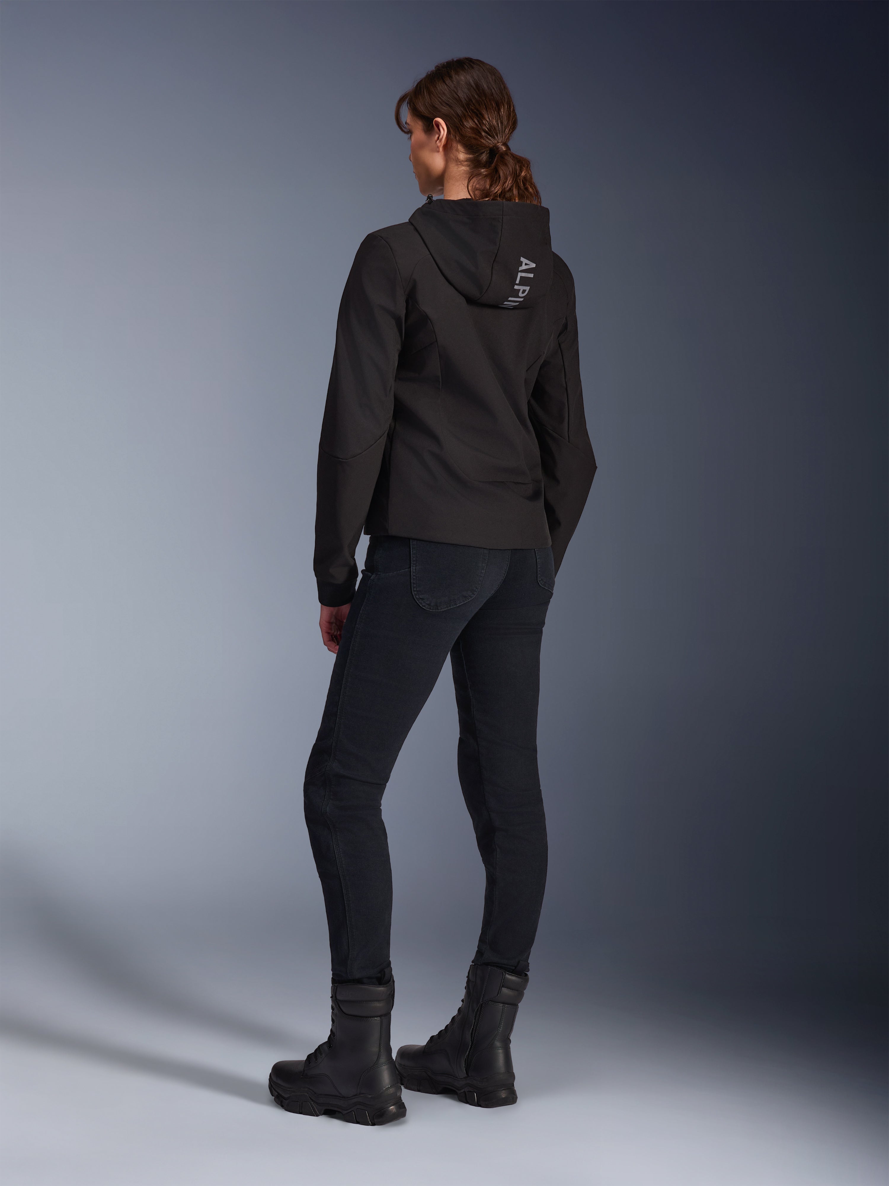 Alpinestars Stella Radium Tech Hoodie, zwarte urban motorhoodie voor dames, achteraanzicht met 'ALPIN'-branding op de capuchon, gecombineerd met een zwarte technische motorbroek en zwarte laarzen, lichtgewicht windbestendige tech shell.