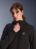 Alpinestars Woman Stella Radium Tech Hoodie, zwart, gedragen door een vrouw die de rits aan de voorkant verstelt, voorzien van een ronde Alpinestars logopatch op de borst, geribde stretch manchetten en een winddichte textielconstructie voor stedelijk rijden.