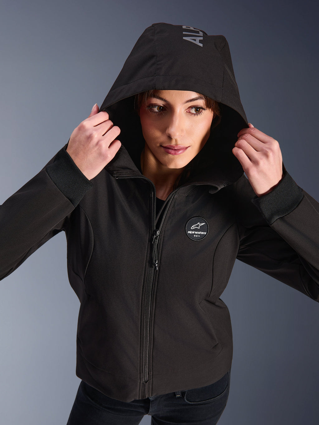 Vrouw die de Alpinestars Stella Radium Tech Hoodie in het zwart draagt, urban motorhoodie met de capuchon omhoog, voorzien van een rond Alpinestars-logo op de borst, geribbelde manchetten en winddichte 3-laagse constructie